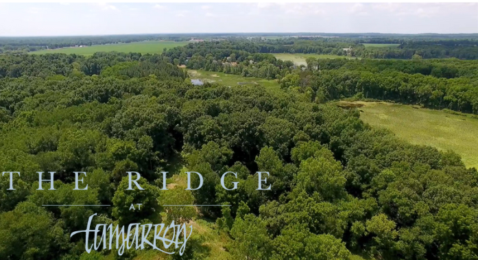 Forested aerial landscape dappled with meadow, marshy lakes, and a blue sky horizon. The logo, reading "The Ridge at Tamarron" sits in grey lettering on the bottom left corner of the image.