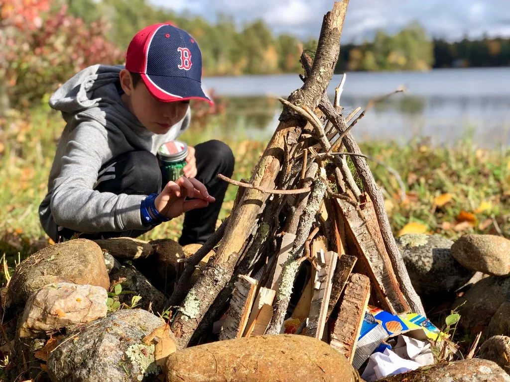 A boy wearing a Boston Red Sox cap, gray hoodie, and black pants, crouches near a fire made of sticks and branches arranged in a teepee shape on a rocky area. There is a lake and trees in the background.