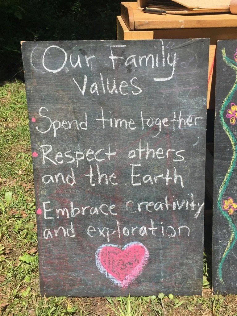 A black chalkboard sign outdoors with handwritten white and pink chalk text displaying family values: "Our Family Values," "Spend time together," "Respect others and the Earth," "Embrace creativity and exploration," and a pink heart at the bottom
