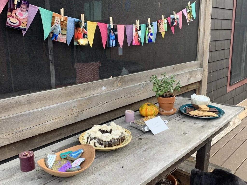 A wooden outdoor table set for a celebration, decorated with a small pumpkin, potted plant, candles, decorated cookies, and various snacks. Colorful photos of children are hanging on a string across a screened porch in the background.