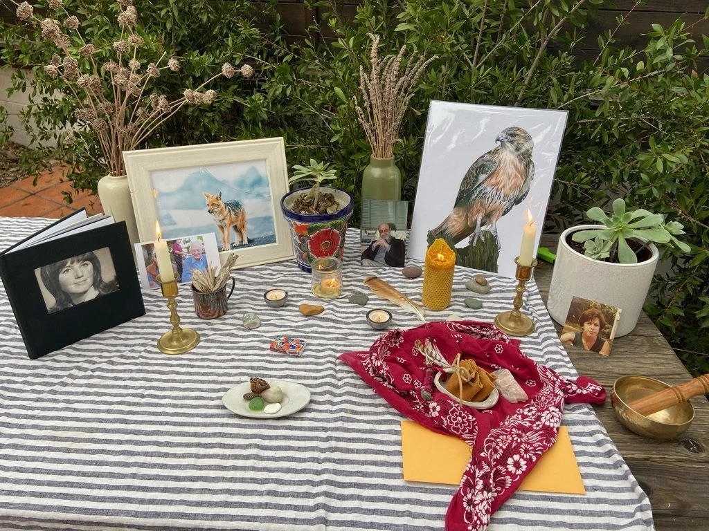 Memorial table with framed photographs, candles, flowers, and personal items, set outdoors on a striped tablecloth surrounded by greenery.
