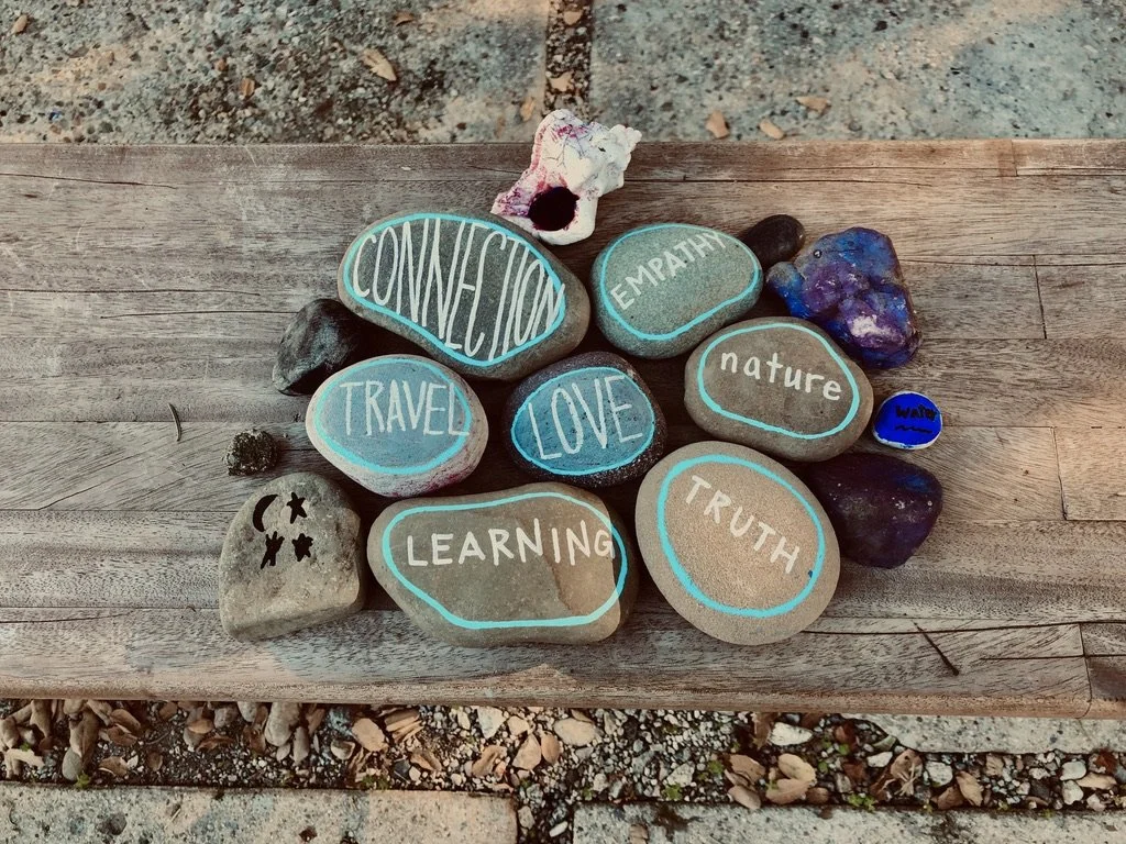 Colorful painted rocks with words on a wooden surface. Words include 'connection,' 'empathy,' 'nature,' 'travel,' 'love,' 'learning,' and 'truth.'