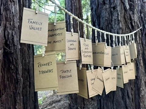 A display of handwritten notes on cardboard cards hanging from a rope tied between trees in a forest, with words like 'Family Values,' 'Rediscover Strengths,' and 'Deeply Feeling'.