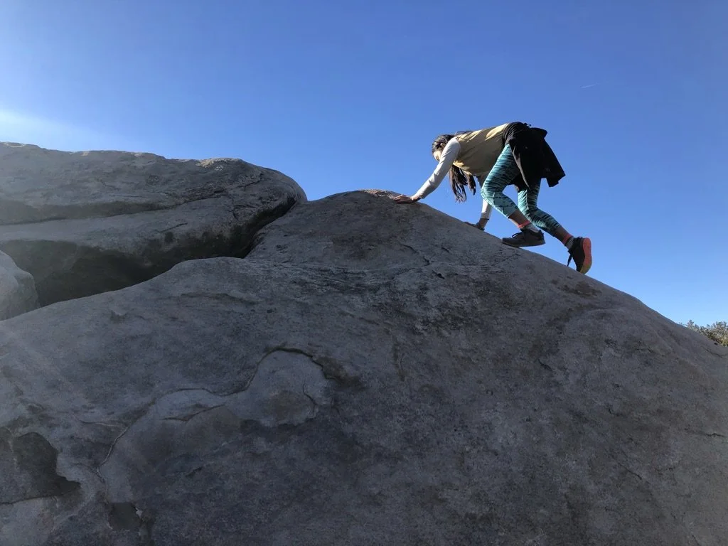 A person wearing a beige vest, teal striped leggings, and black shoes is climbing up a large rock in an outdoor setting with blue sky in the background.