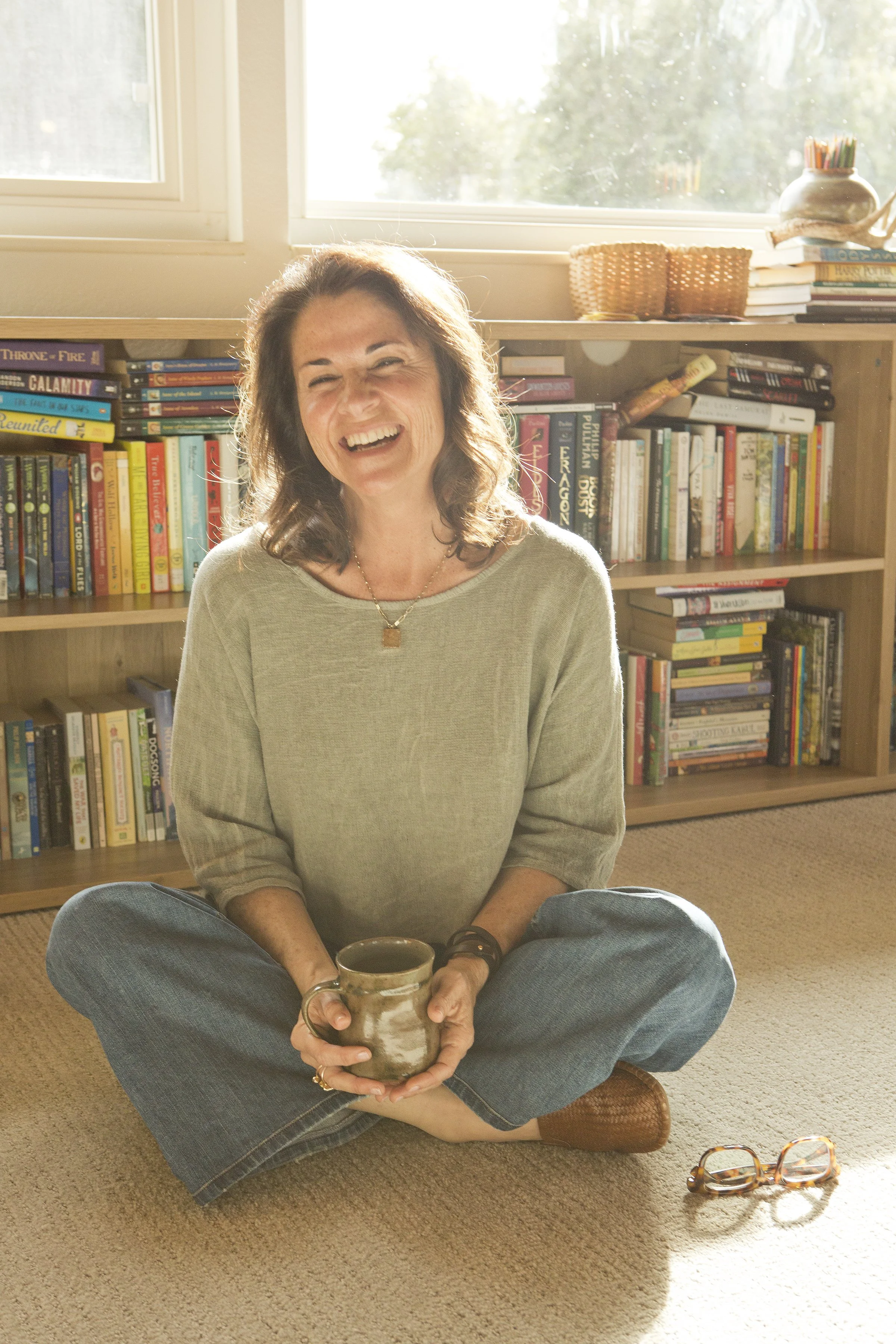A woman sitting cross-legged on a beige carpet, smiling and holding a mug, in front of a wooden bookshelf filled with books. There is sunlight coming in through a large window behind her.