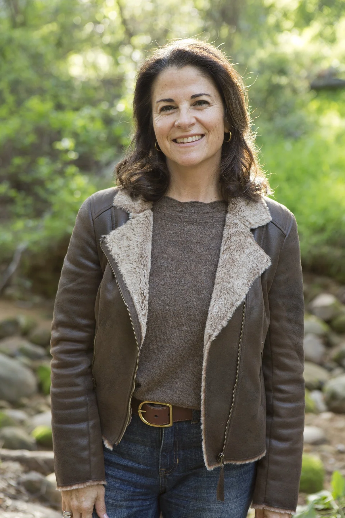 A woman with dark brown hair smiles outdoors in a forested area, wearing a brown leather jacket with a shearling collar, a grey sweater, and blue jeans.