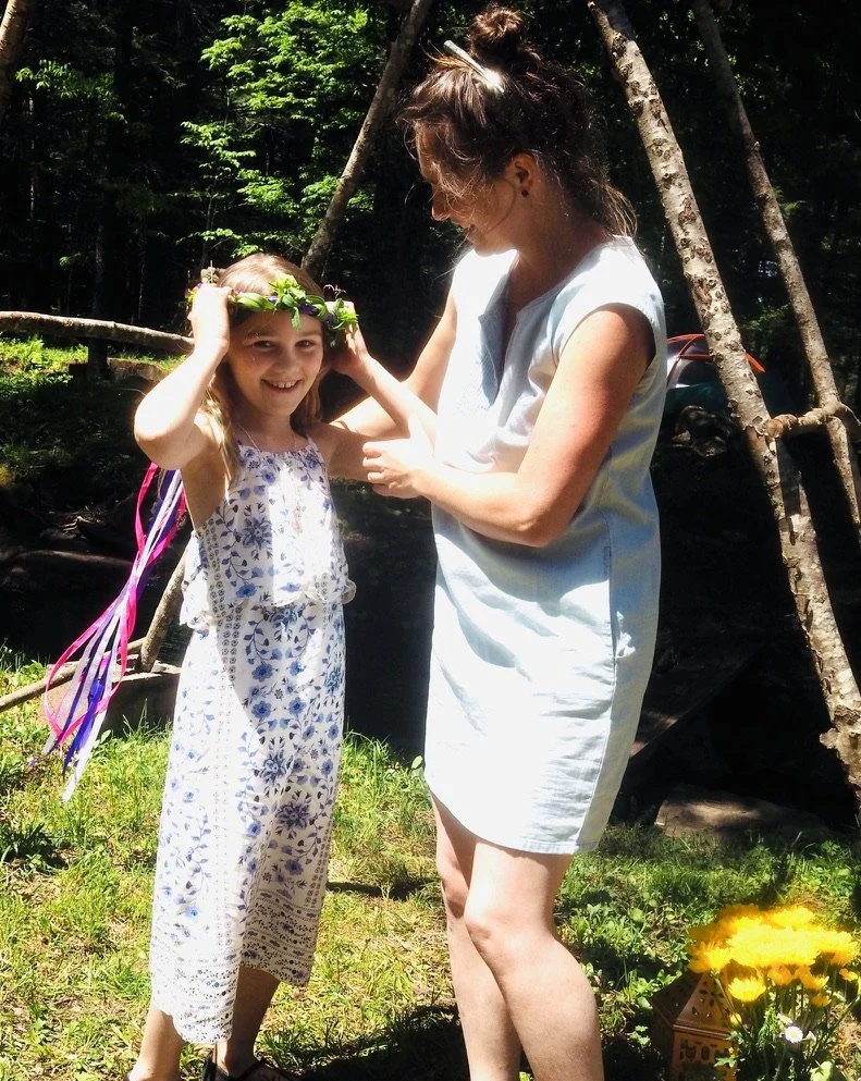 A woman and a girl outdoors, the girl wearing a floral dress and a leafy crown, smiling as the woman helps adjust the crown. They are in a wooded area with trees, grass, and a decorated flowerpot in the foreground.