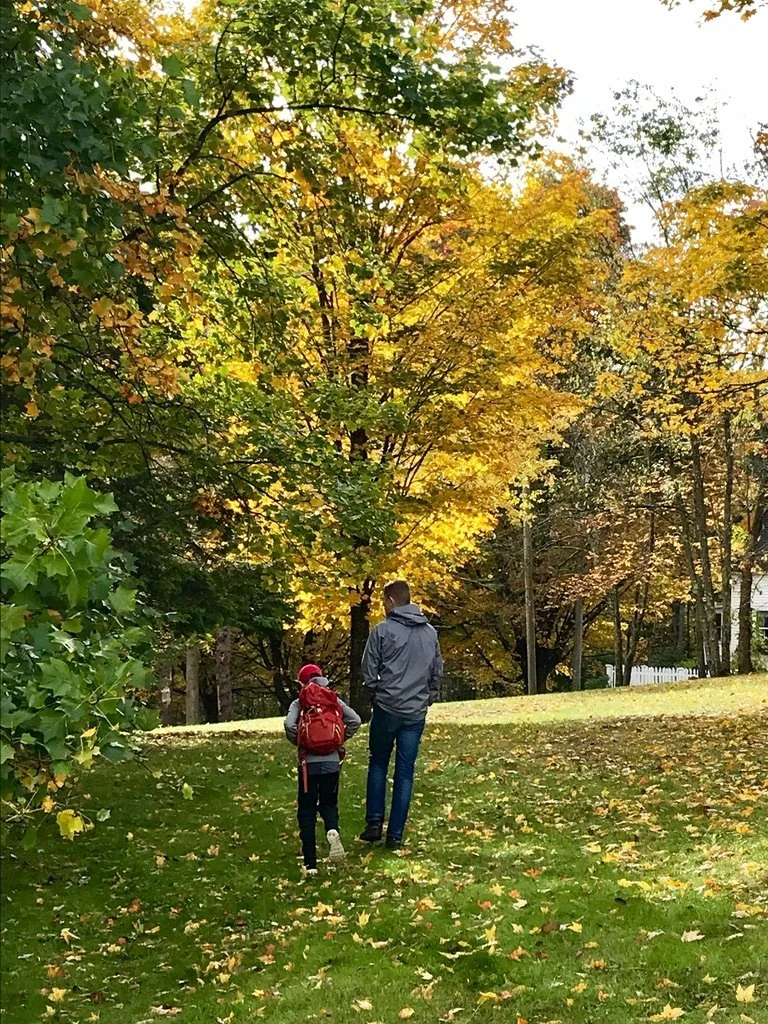 A man and a child walking in a park with green and yellow trees during autumn. The child is wearing a red backpack and red hat, and the man is wearing a gray jacket.