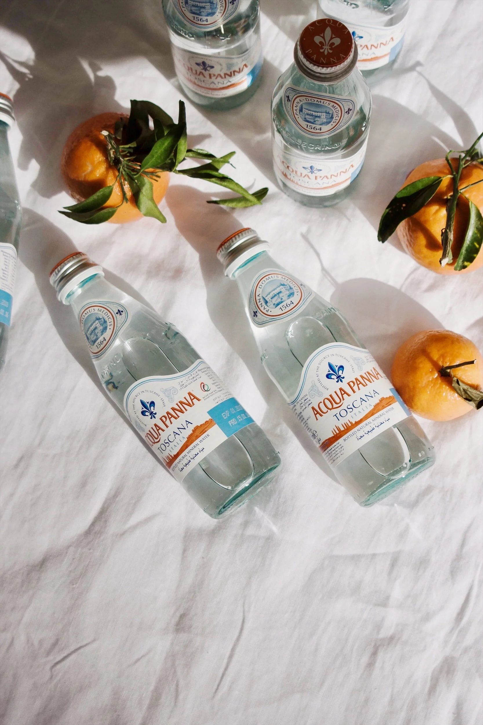 Glass bottles of water and oranges arranged on a white sheet