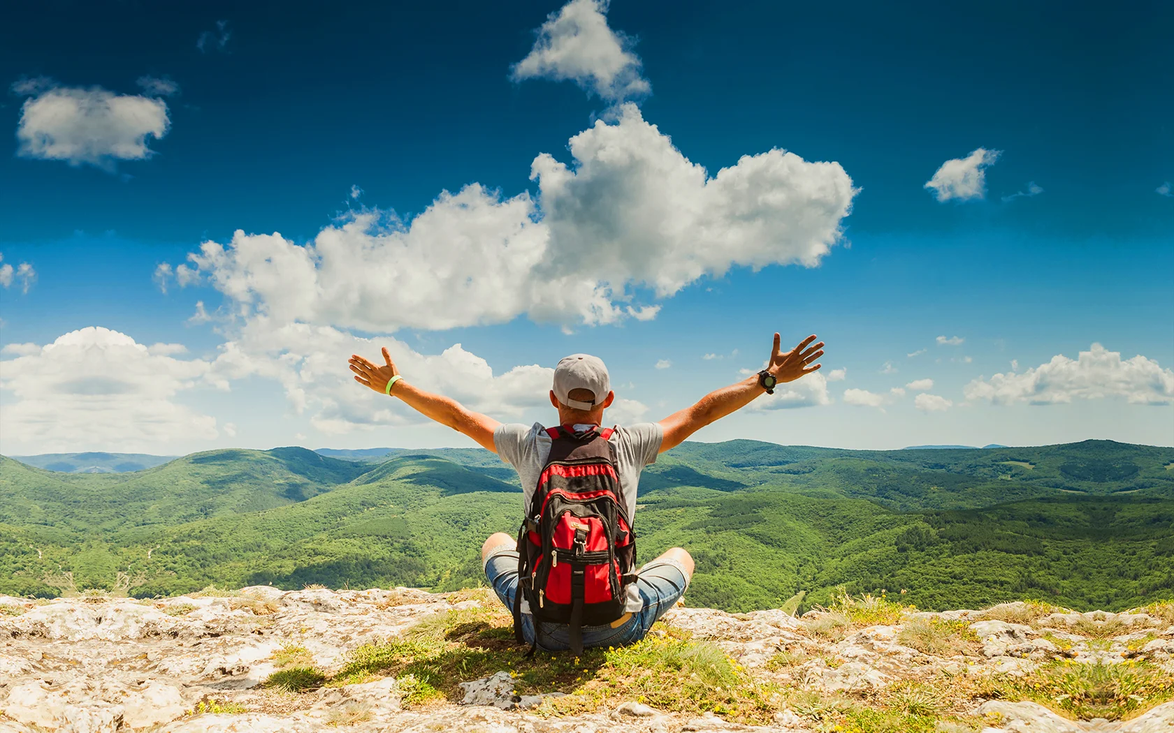 A man sitting on a rocky hilltop with arms outstretched, overlooking a green mountain landscape under a partly cloudy blue sky.