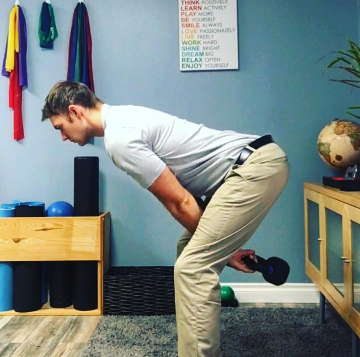 Young man performing a bent-over row exercise with a dumbbell in a home gym. He is in a zigzag posture, holding the dumbbell with his right hand, wearing a gray T-shirt and khaki pants. The room has a blue wall with a motivational poster, colorful resistance bands hanging on the wall, a wooden shelf with exercise equipment, and a wooden cabinet with a potted plant on top.
