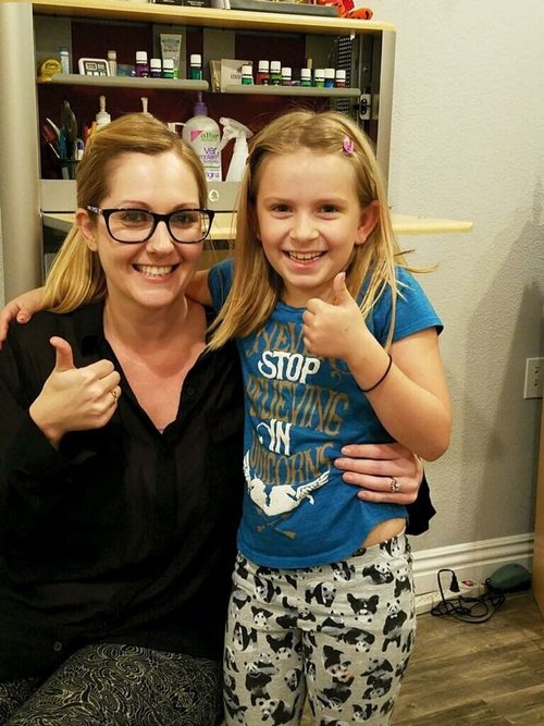 A woman and young girl smiling and giving thumbs up indoors. The woman has glasses and blonde hair, and the girl has blonde hair with a pink hair clip, wearing a blue T-shirt and patterned pants.