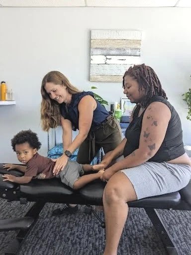 A woman receiving a massage from a female massage therapist while a young child rests on a massage table.