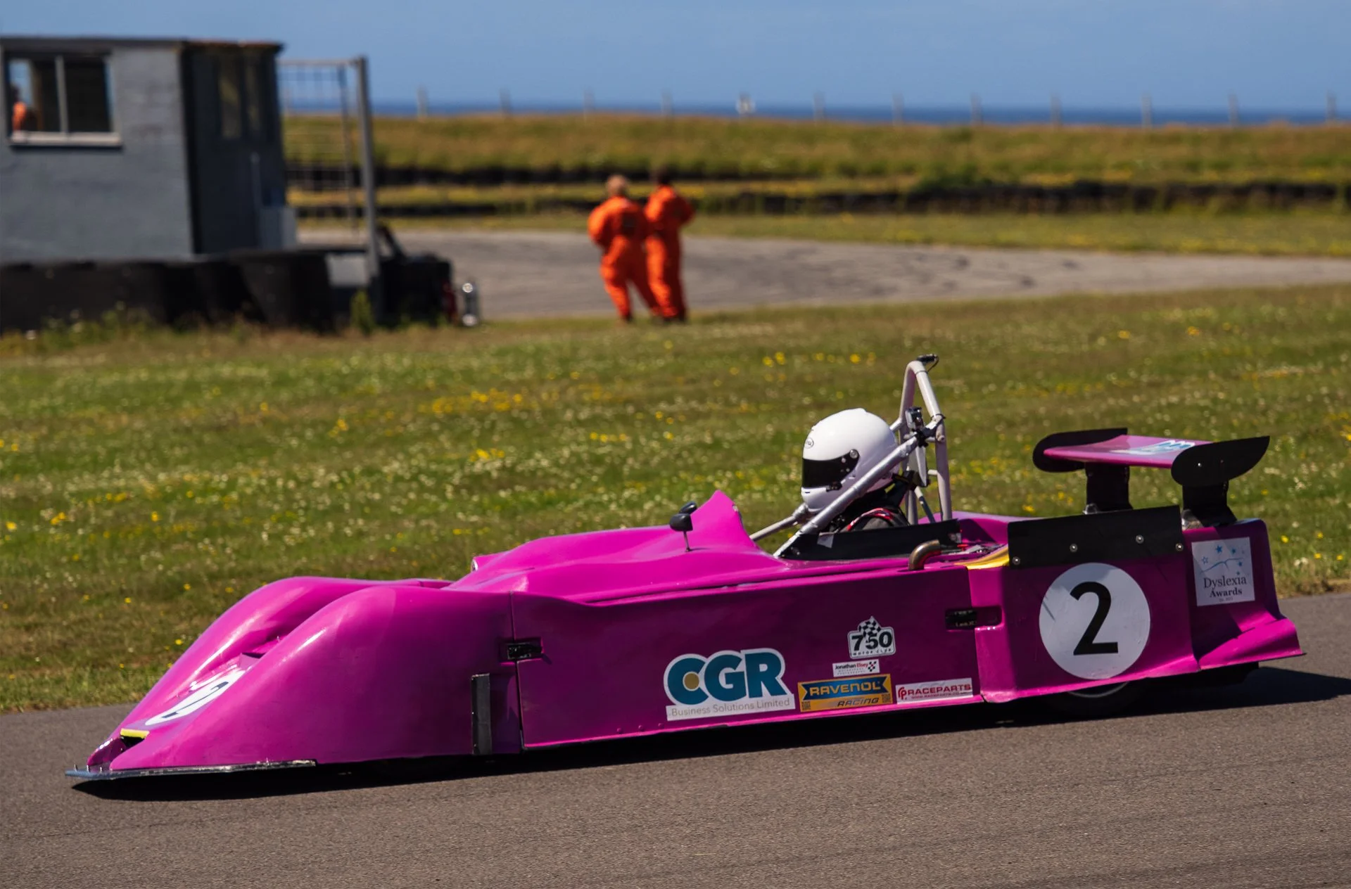 Close-up of a pink race car with racing stickers and the number 2, on a paved track with two people in orange suits in the background near the grass, and an outbuilding or trailer to the left.