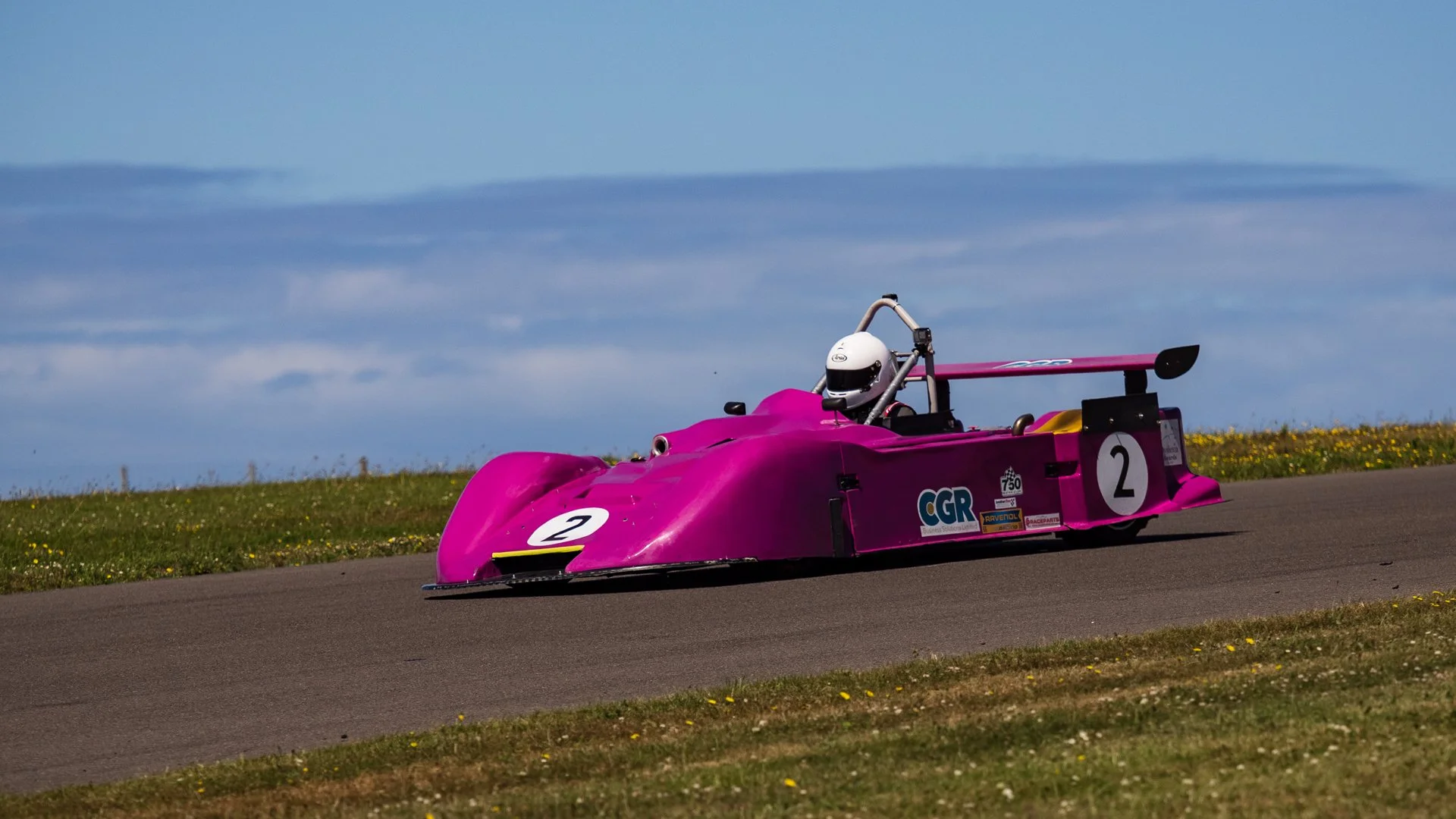 A pink racing car with a white number 2 on the front and side, driven by a person wearing a white helmet, moving on a paved race track with a grassy field and blue sky in the background.
