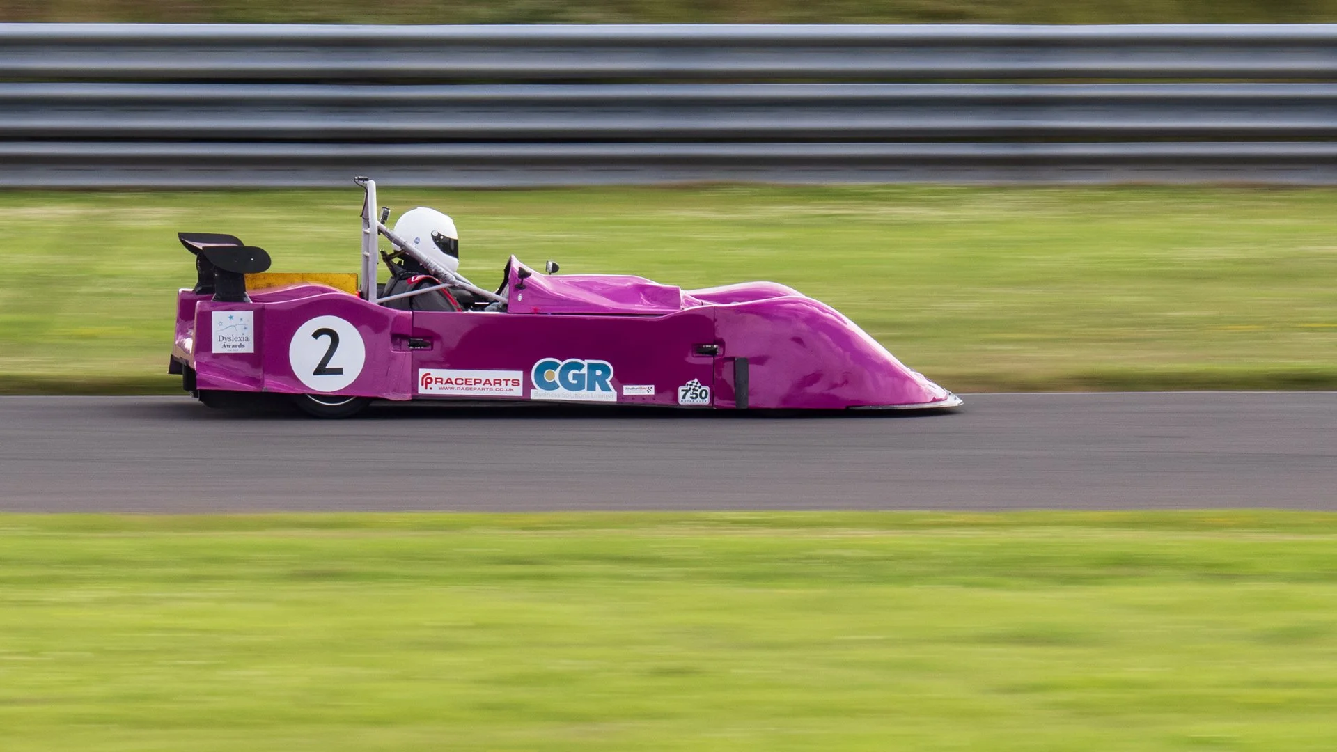 Pink racing car with the number 2 on the side, driven by a driver in a white helmet, speeding on a race track, with a grassy area and a metal barrier in the background.
