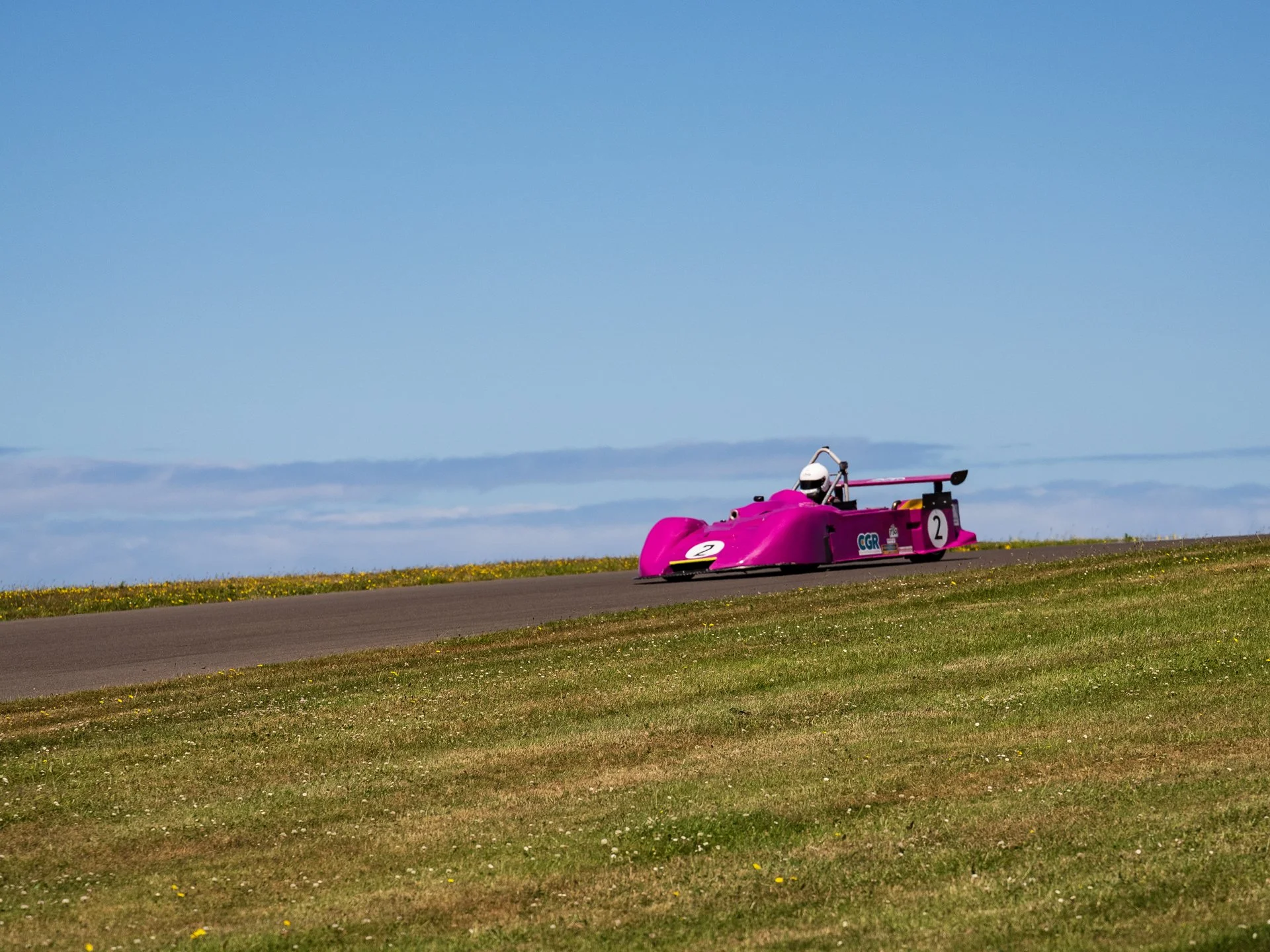 A pink race car on a race track with grass on the sides and a clear blue sky in the background.