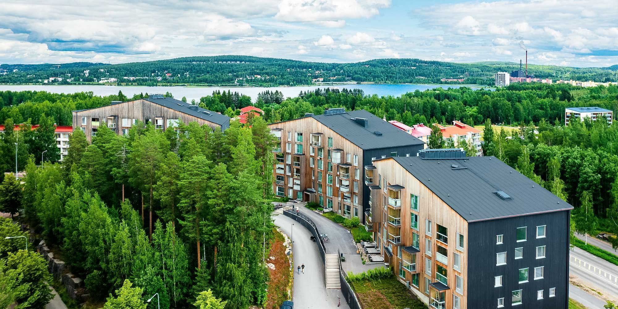 Dronekuva kaupunkimaisemasta kiinteistöineen Jyväskylän Kuokkalasta, kuvassa Puukuokka-nimiset puiset kerrostalot. Vihreitä puita, taustalla sininen Jyväsjärvi.
