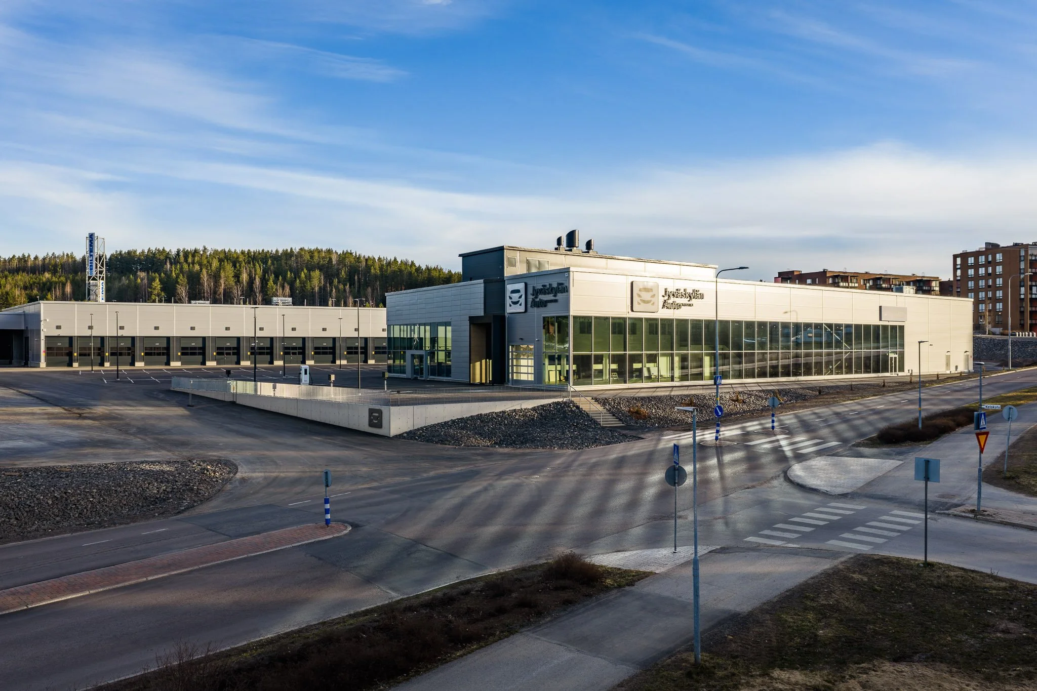 Jyväskylän Autotarvikkeen uudet rakennukset Palanderinkadulla kuvattuna donella aamuauringossa.Etualalla automyynnin rakennus ja taustalla autohuollon rakennus. Taivas on seesteisen sininen, auringonvalo heijastuu tiehen tehden siihen raitoja.