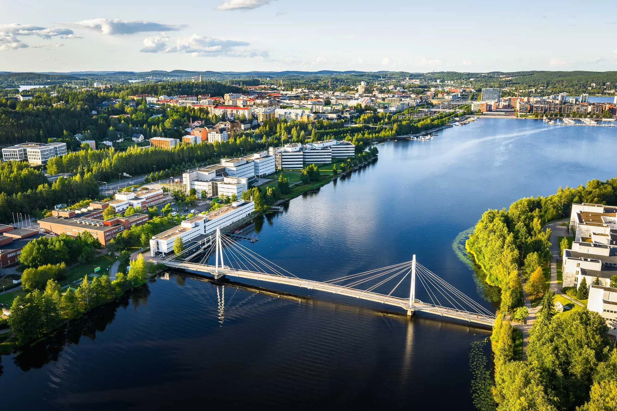Dronekuva Jyväskylästä Ylistön kevyen liikenteen sillan luota, kuvassa Jyväsjärvi ja kaupungin keskustaa kesäisessä iltavalossa, keskellä kuvaa hotelli Alba, Yliopiston Agora ja Kielon yritystalot
