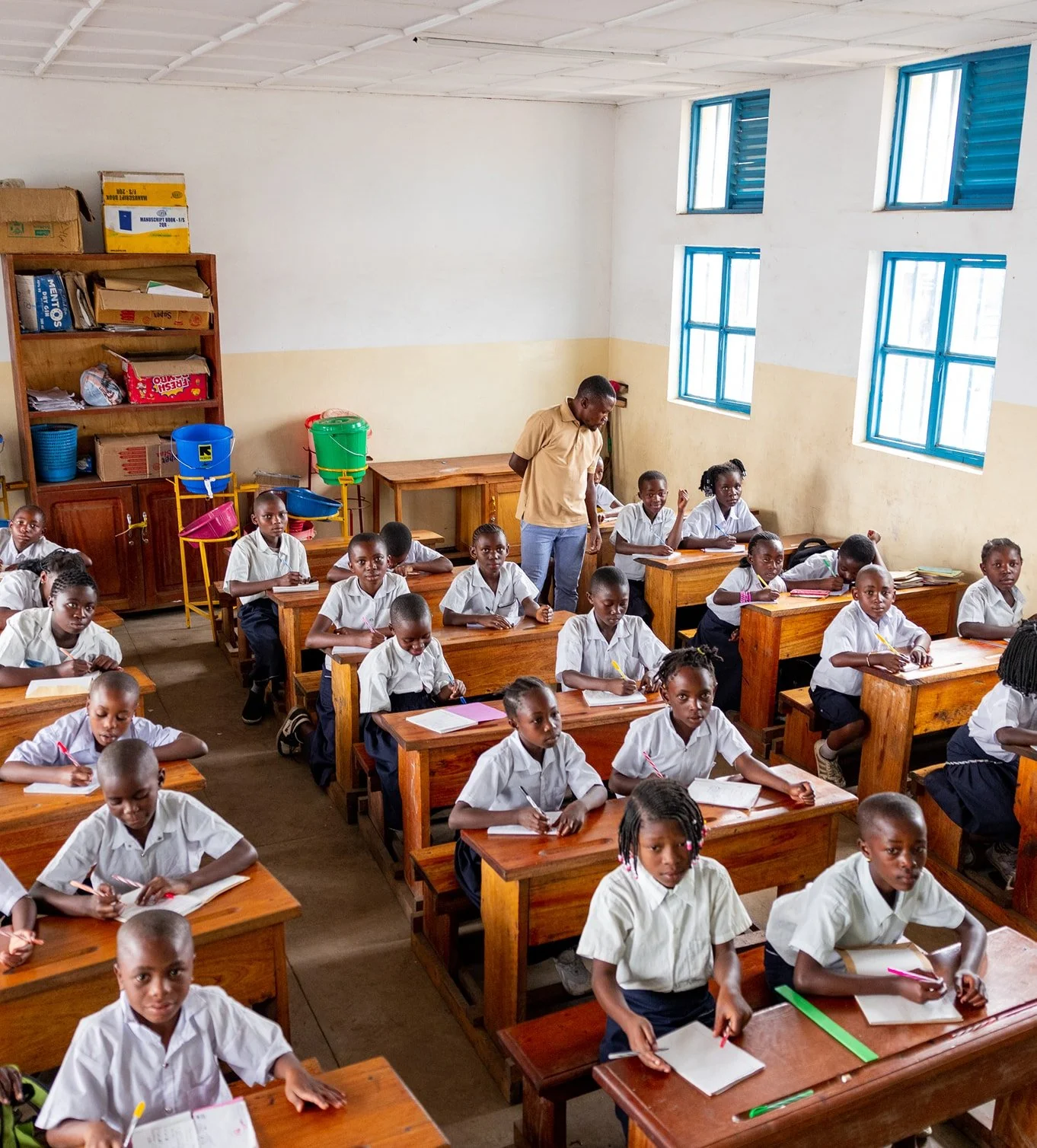 Students sit at wooden desks in a classroom, writing in notebooks while a teacher stands among them, guiding their work in a bright, orderly learning environment.