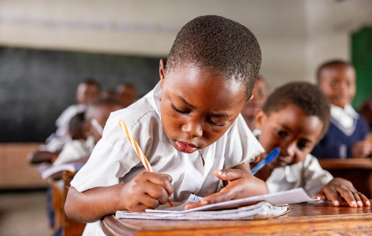 Young students sit at wooden desks in a classroom, with one child in the foreground concentrating on writing in a notebook while others work quietly behind.