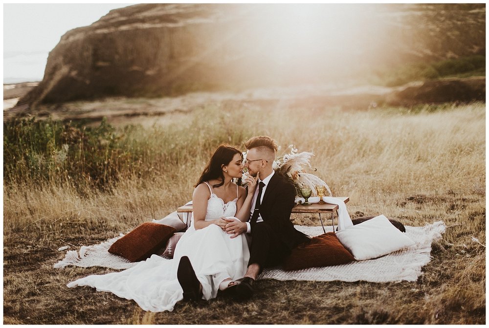 a good looking young married couple sit at their picnic-style meal in the desert, the sun setting behind them