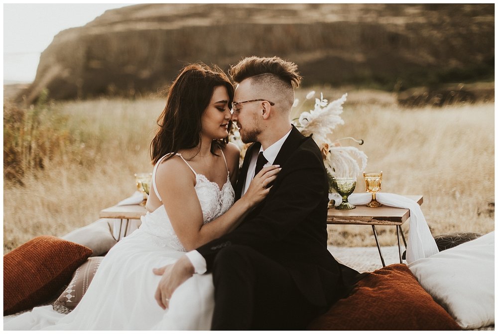 a bride and groom sit at their boho picnic in the desert and snuggle up to one another