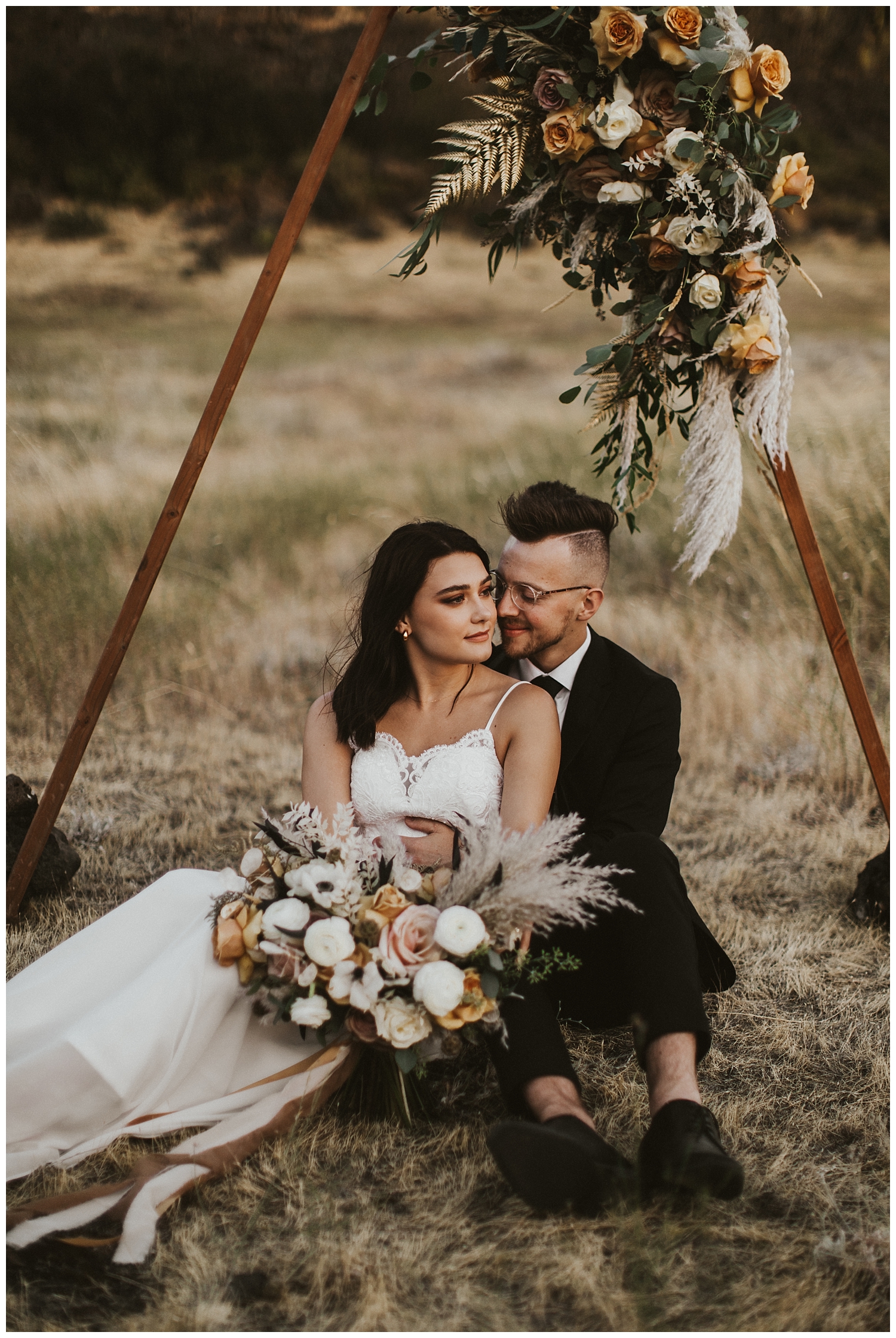 a boho bride and groom sit together under their wooden triangle arch in the desert