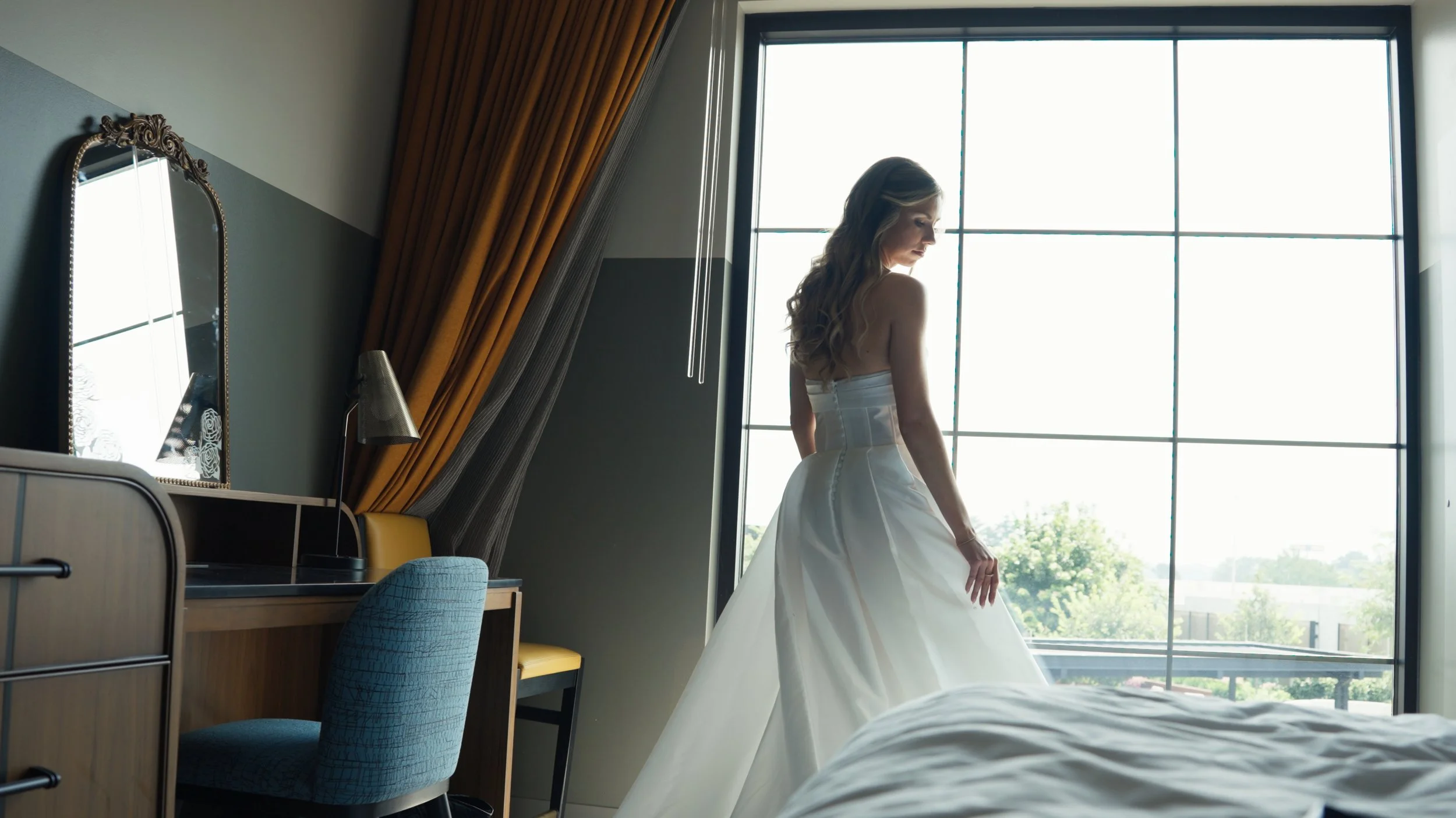 A woman in a white wedding dress standing near a large window in a hotel room, with sunlight illuminating her and her surroundings.