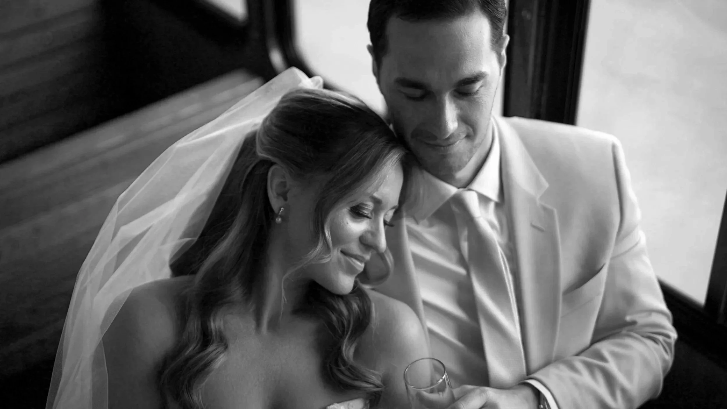 A black-and-white photo of a bride and groom at their wedding, sitting close with their heads touching, smiling gently. The bride is wearing a veil and earrings, and the groom is dressed in a white suit. The bride holds a glass of champagne.