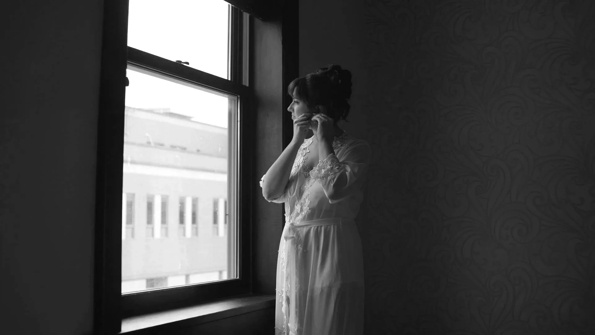 A woman in a lace robe looking out of a window, adjusting her earrings, in a dimly lit room.