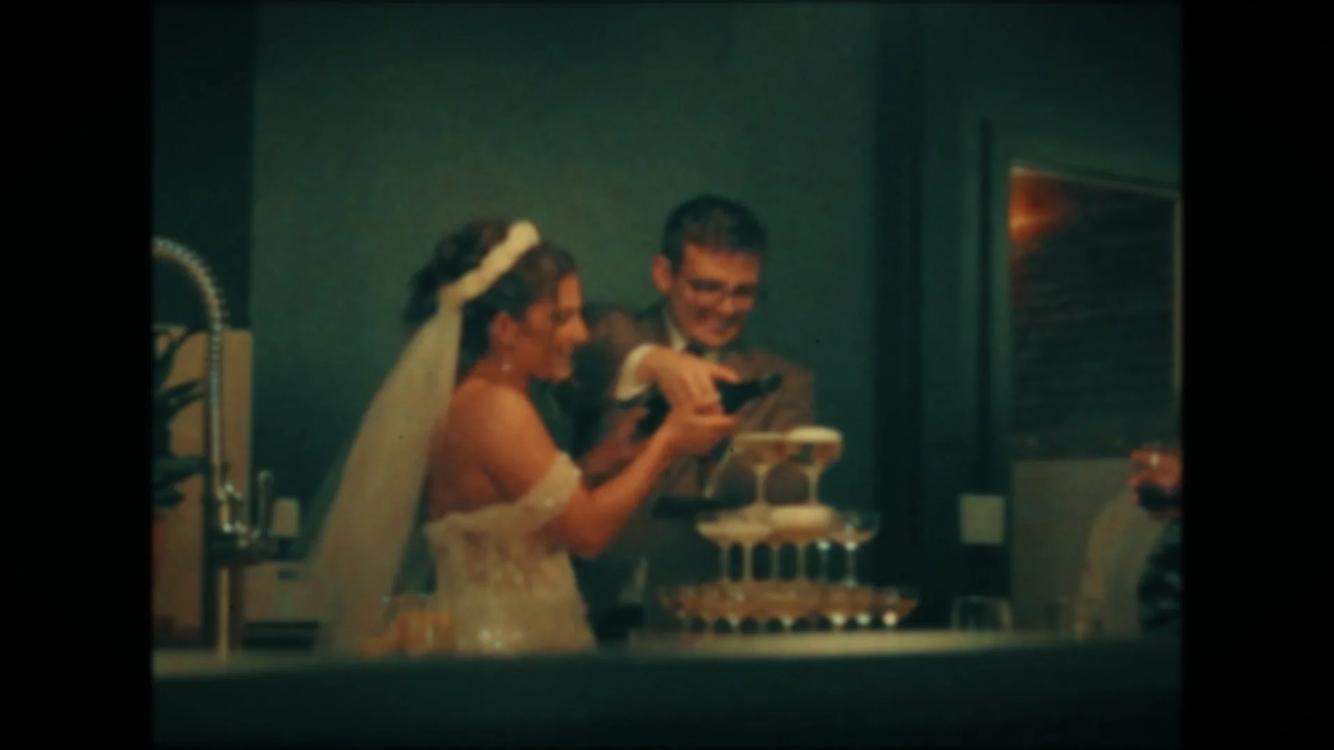 A bride and groom in wedding attire sharing a joyful moment looking at something together, possibly a cake, with a cake on a table in front of them.