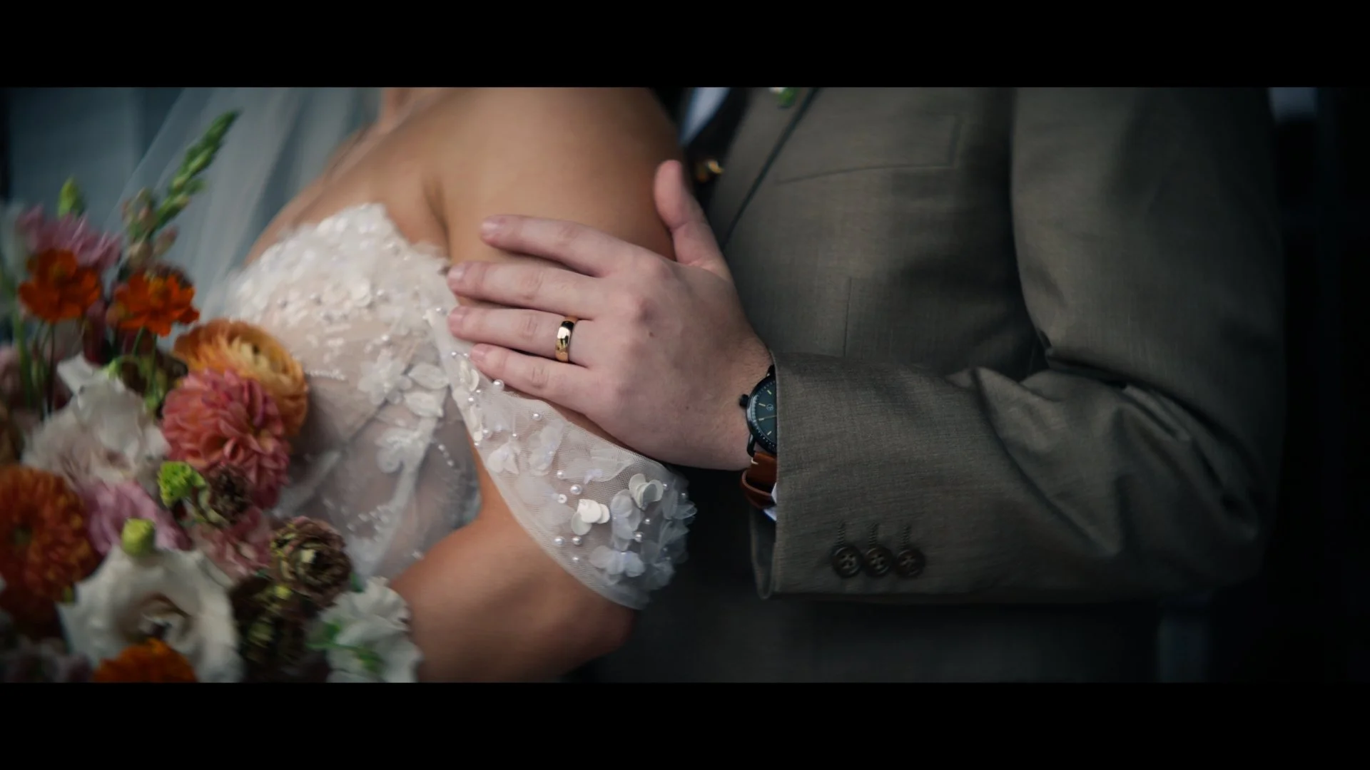 Close-up of a bride and groom holding hands during a wedding, with the groom's hand on the bride's shoulder showing wedding rings, and a bouquet of flowers nearby.
