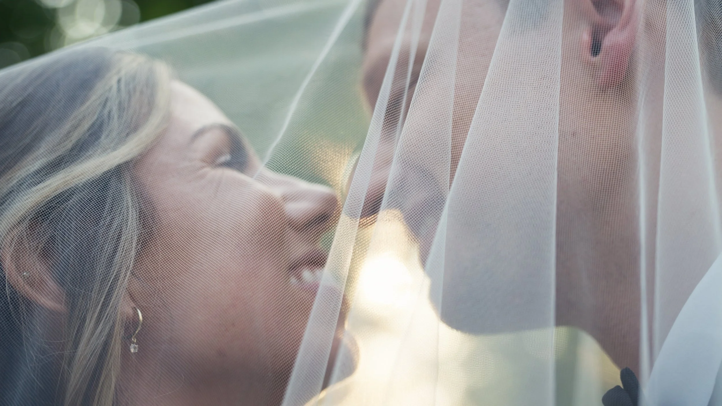 Bride in a wedding dress smiling behind a sheer veil with someone adjusting it, outdoors during sunset.