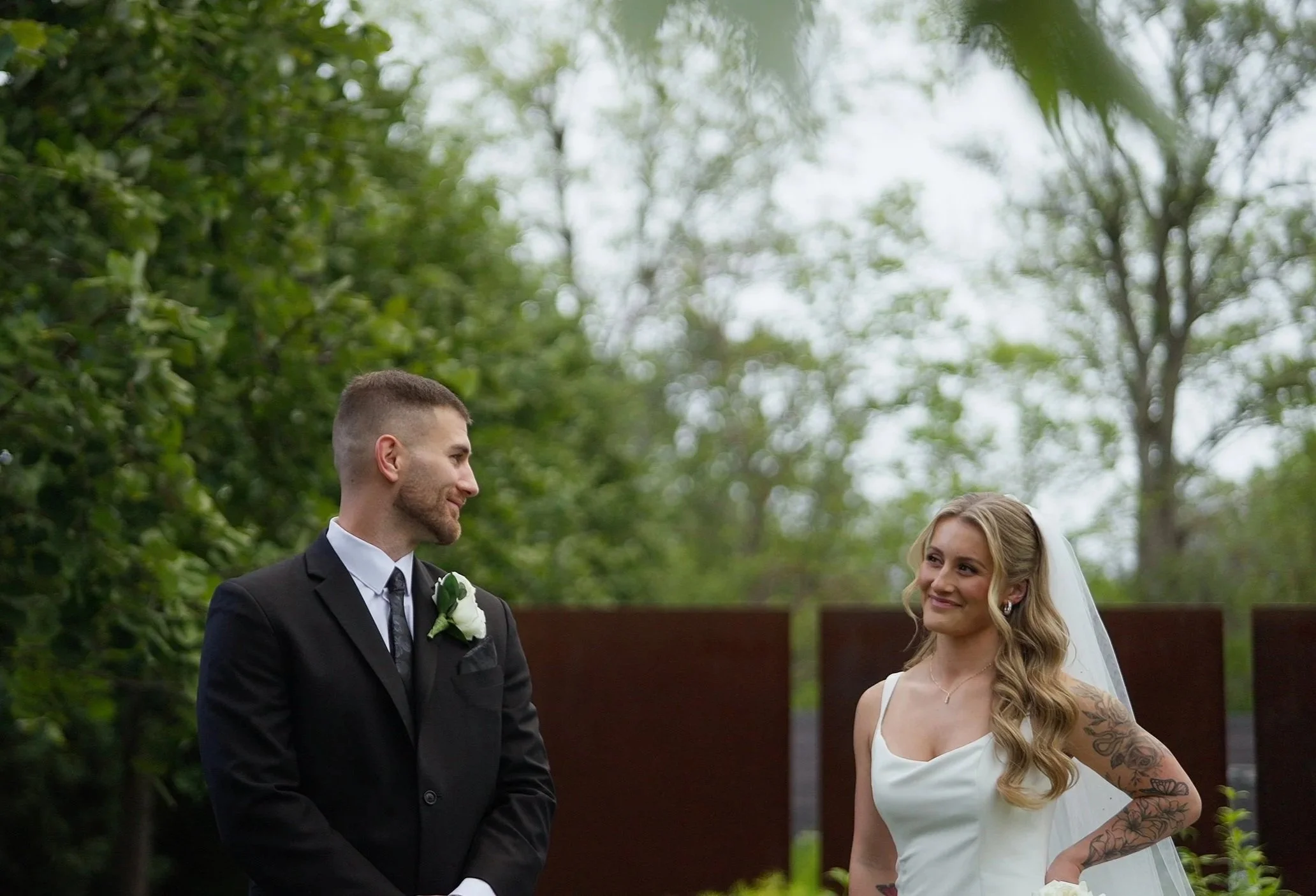 A bride and groom standing outdoors during their wedding ceremony, smiling at each other, with lush green trees in the background.