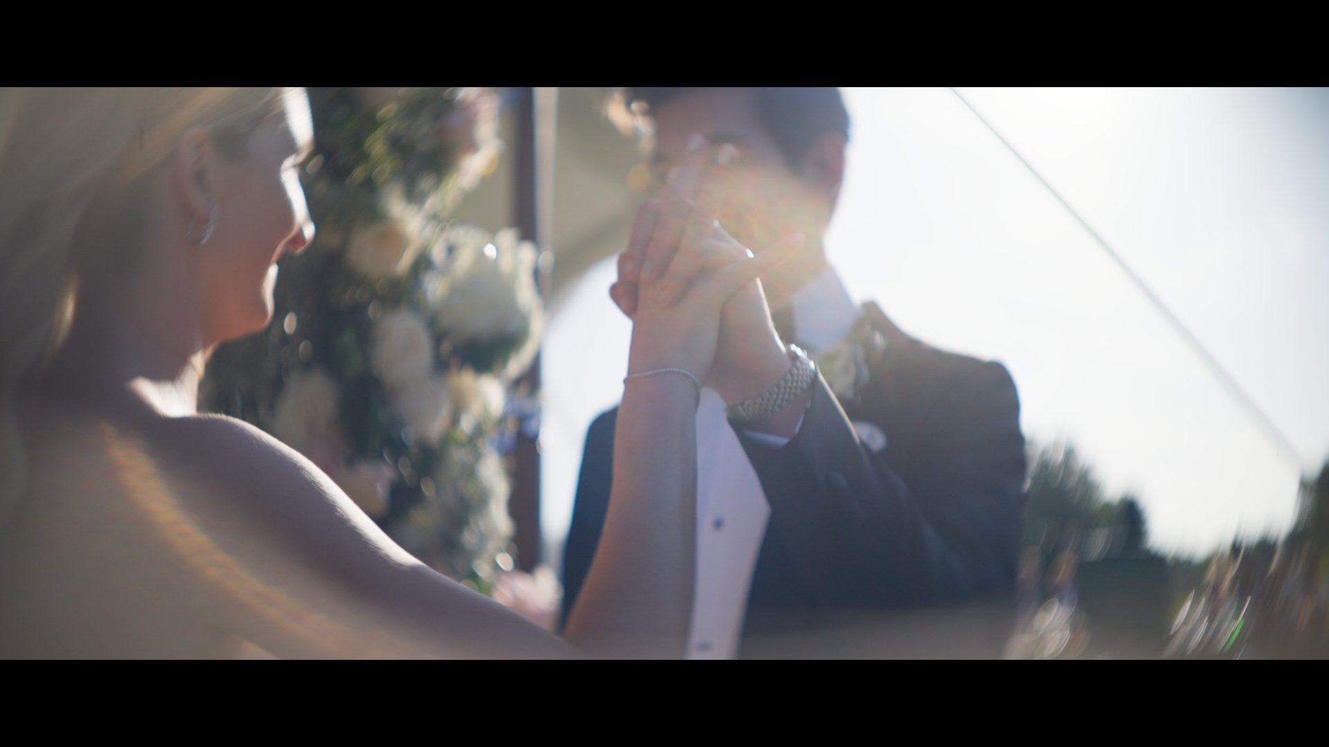 A couple holding hands and smiling at each other at a wedding ceremony outdoors, with sunlight shining from behind them.