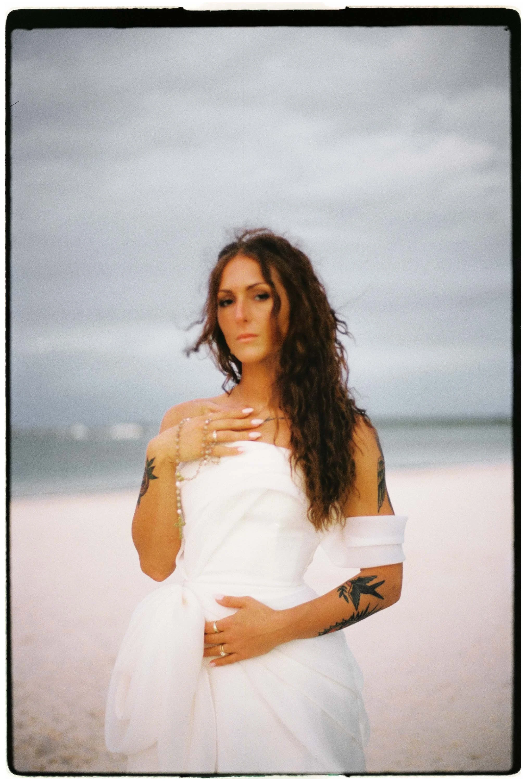 Bride posing as the Boticelli's Venus with the beach and the ocean in the background. She is wearing white and holding a golden crucifix