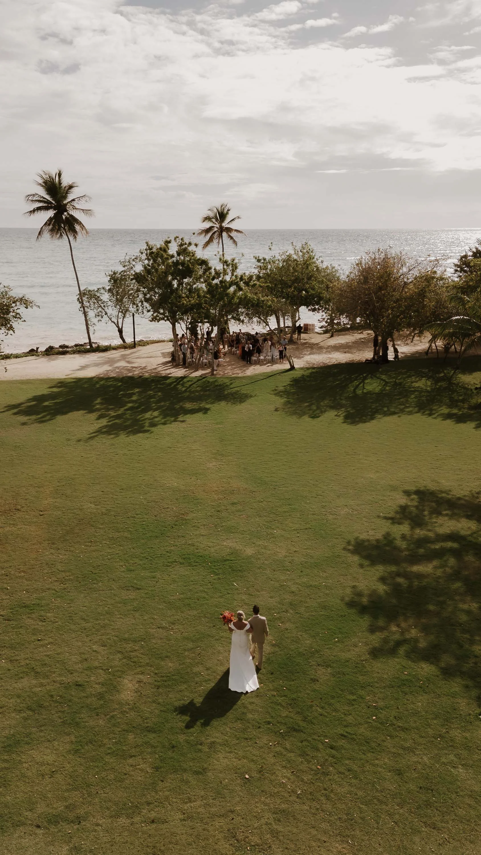 Drone photo of the bride walking towards the ceremony, while the guest and the groom wait for her. The shining sea is in the background and the sun is out.