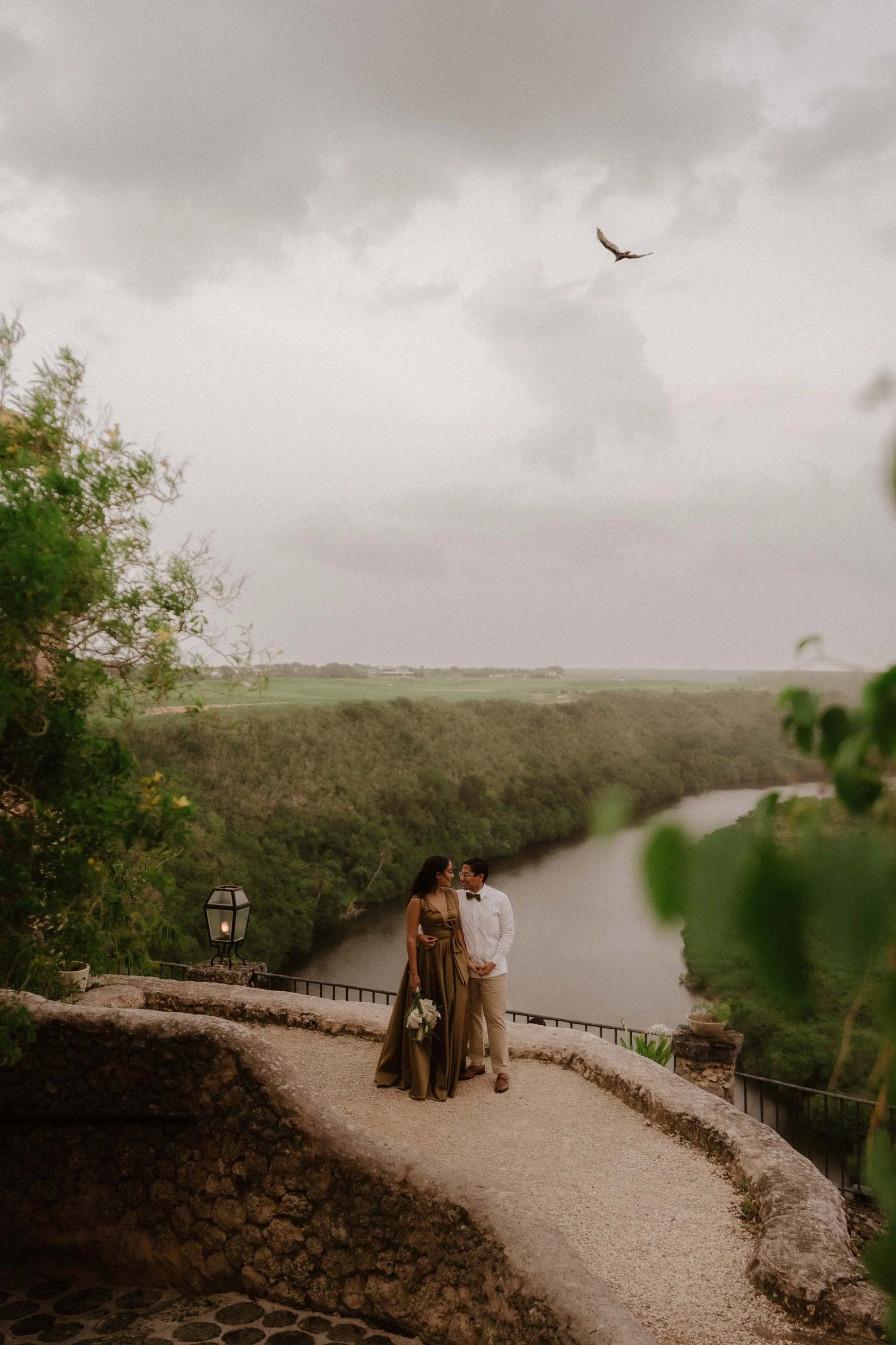 Bride and groom posing for the camera, behind them stands the rio chavon and a bird croses over their head during the sunset