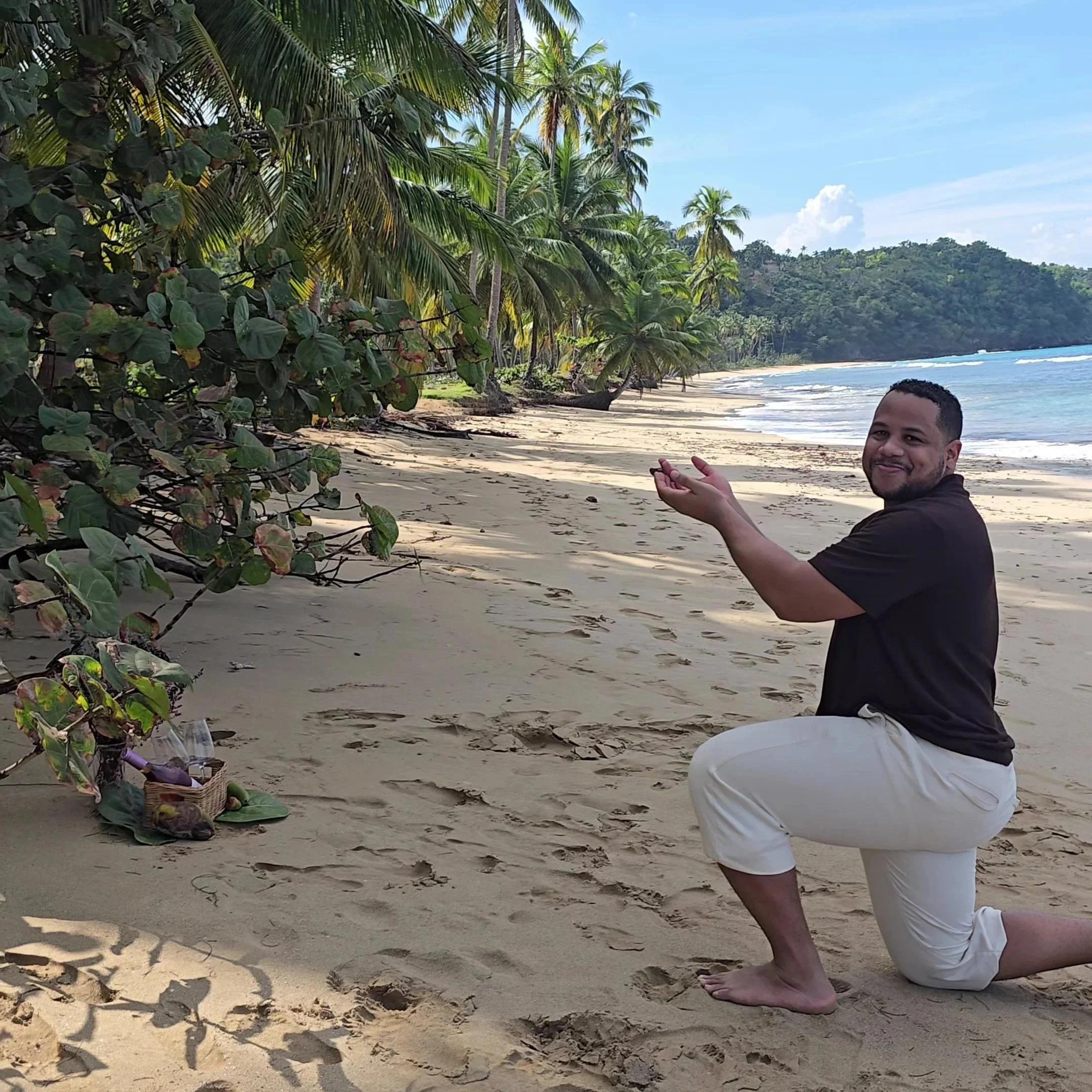 Photographer kneeling down showning to the groom how to propose and how to kneel down for the proposal. At the background the forest and the beach in Samana