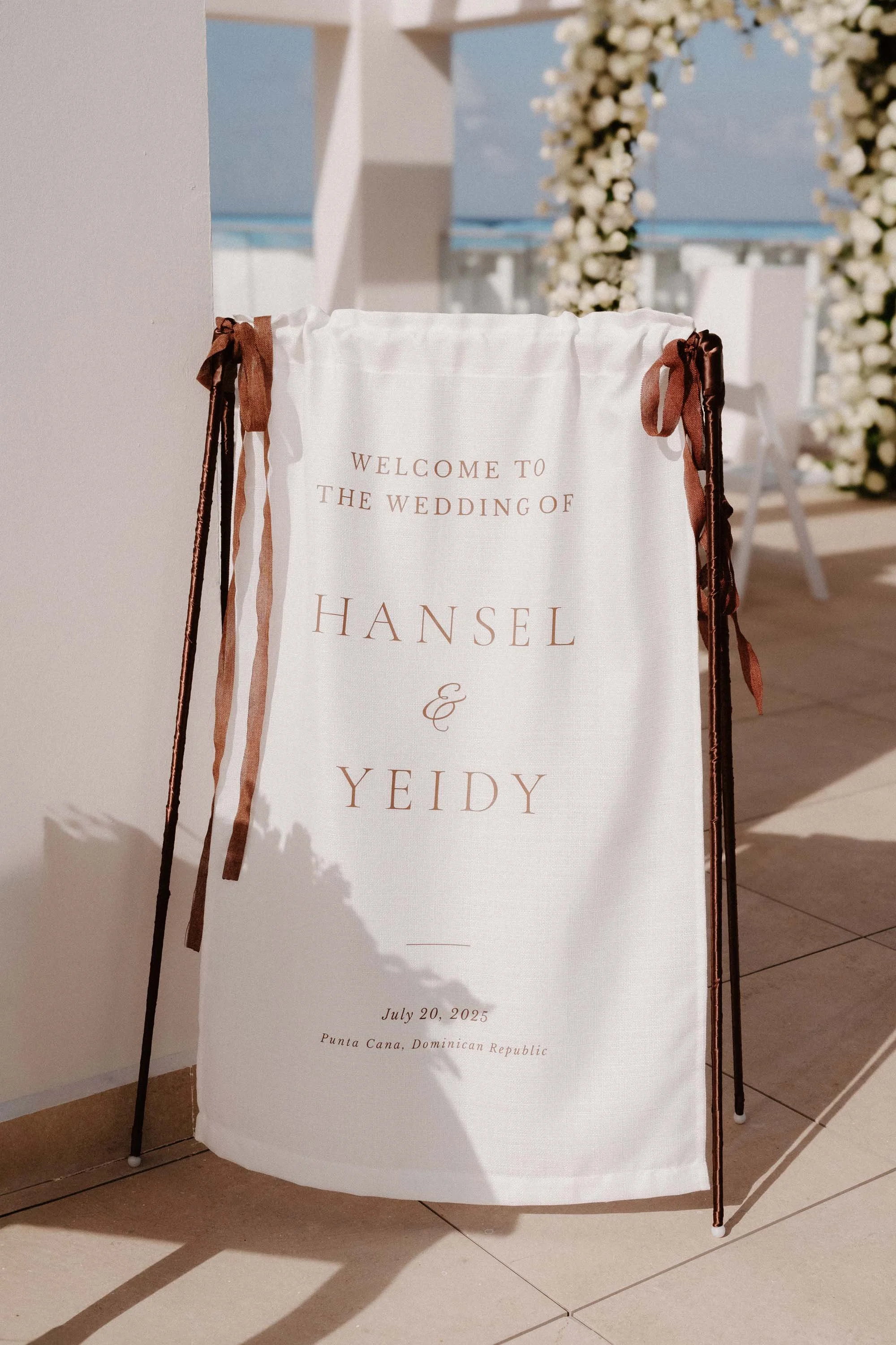 An elegant white fabric wedding welcome sign tied with warm brown ribbons, softly illuminated by the afternoon sun before an intimate destination wedding in Cap Cana Punta Cana