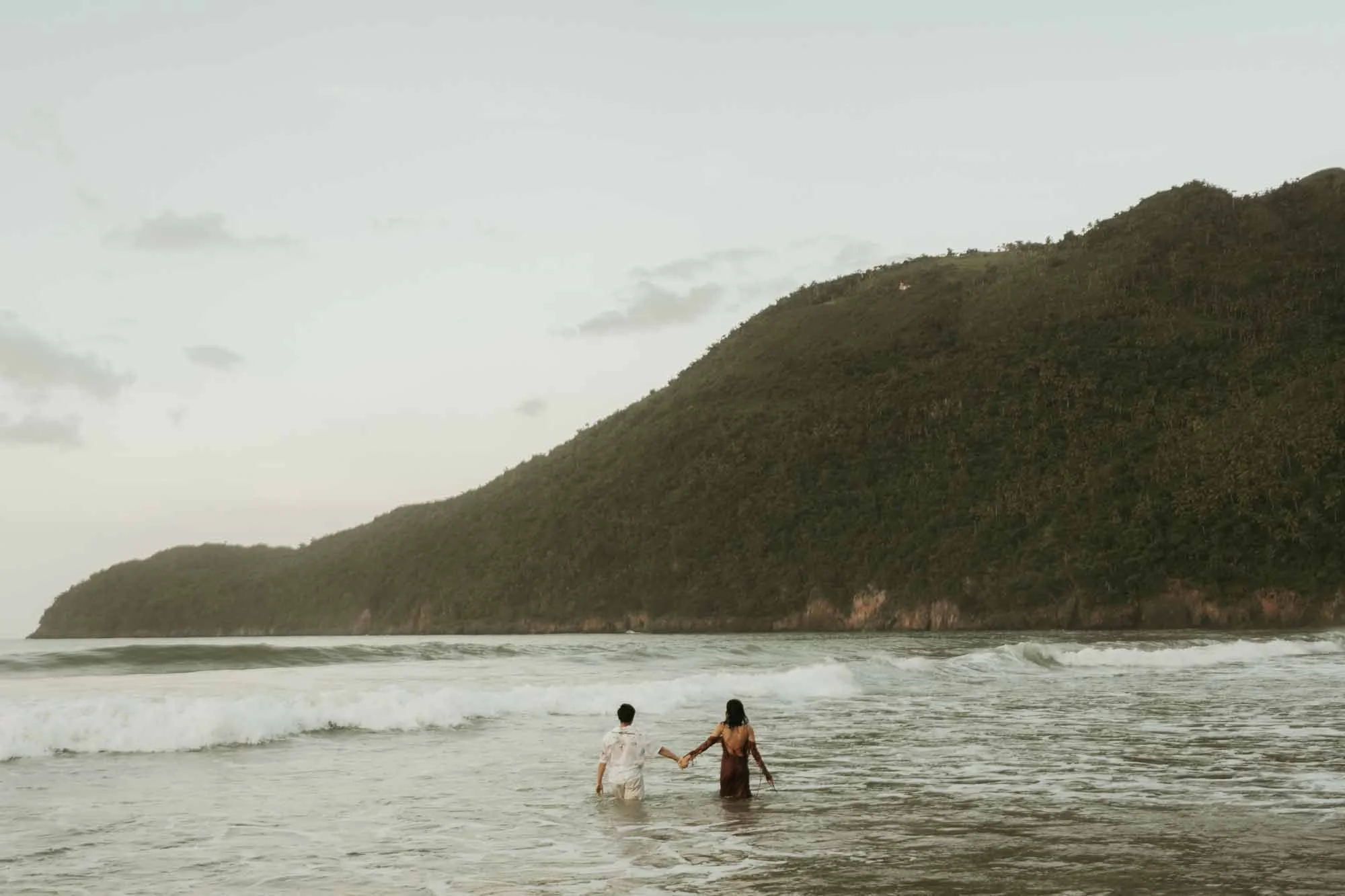 Couples holding hands in an engagement photoshoot in Dominican Republic. They are inside the water and at the background you can see the mountains and the waves.