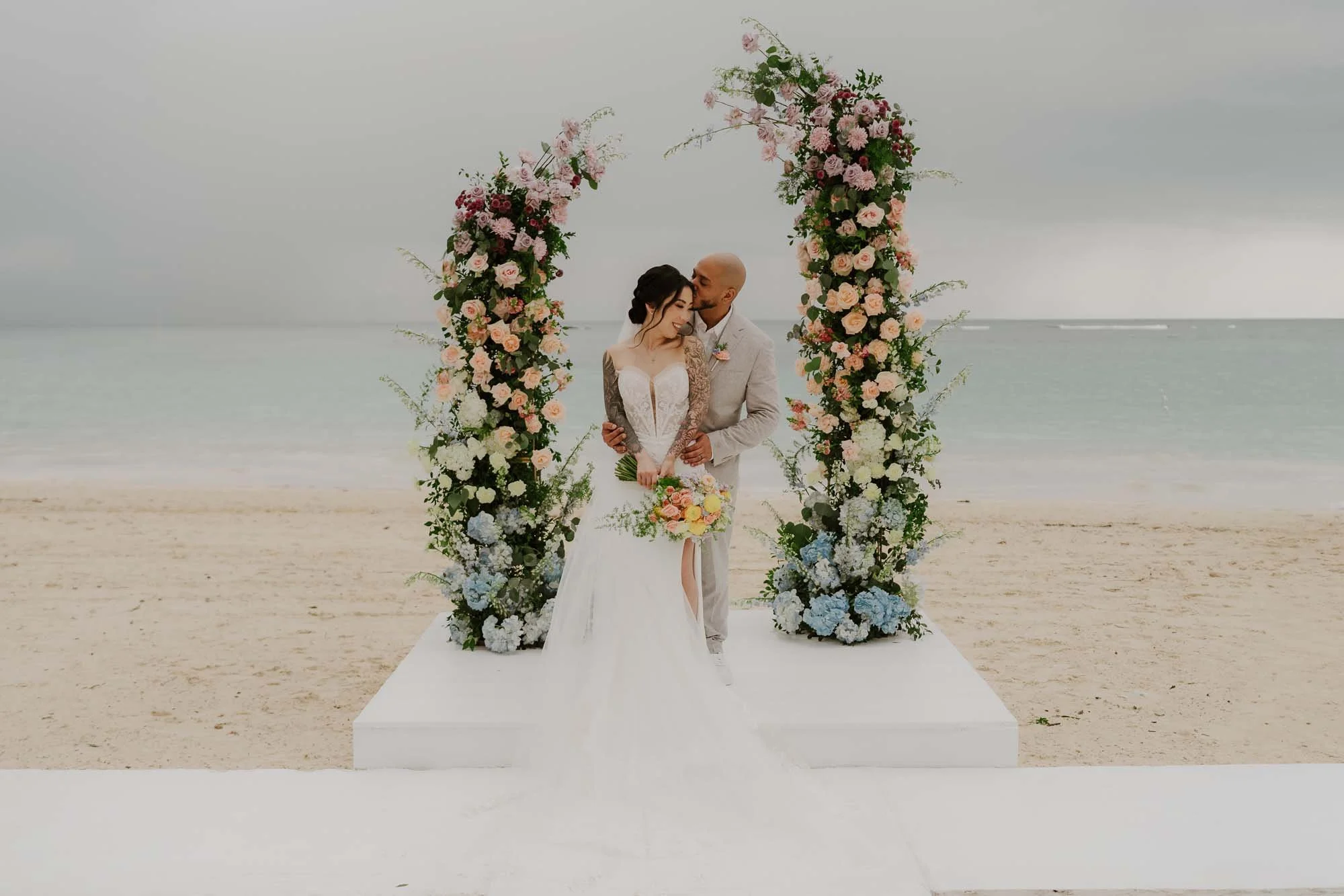 Couple holding hands under an arch full of flowers, in the background you can see the Punta Cana beach