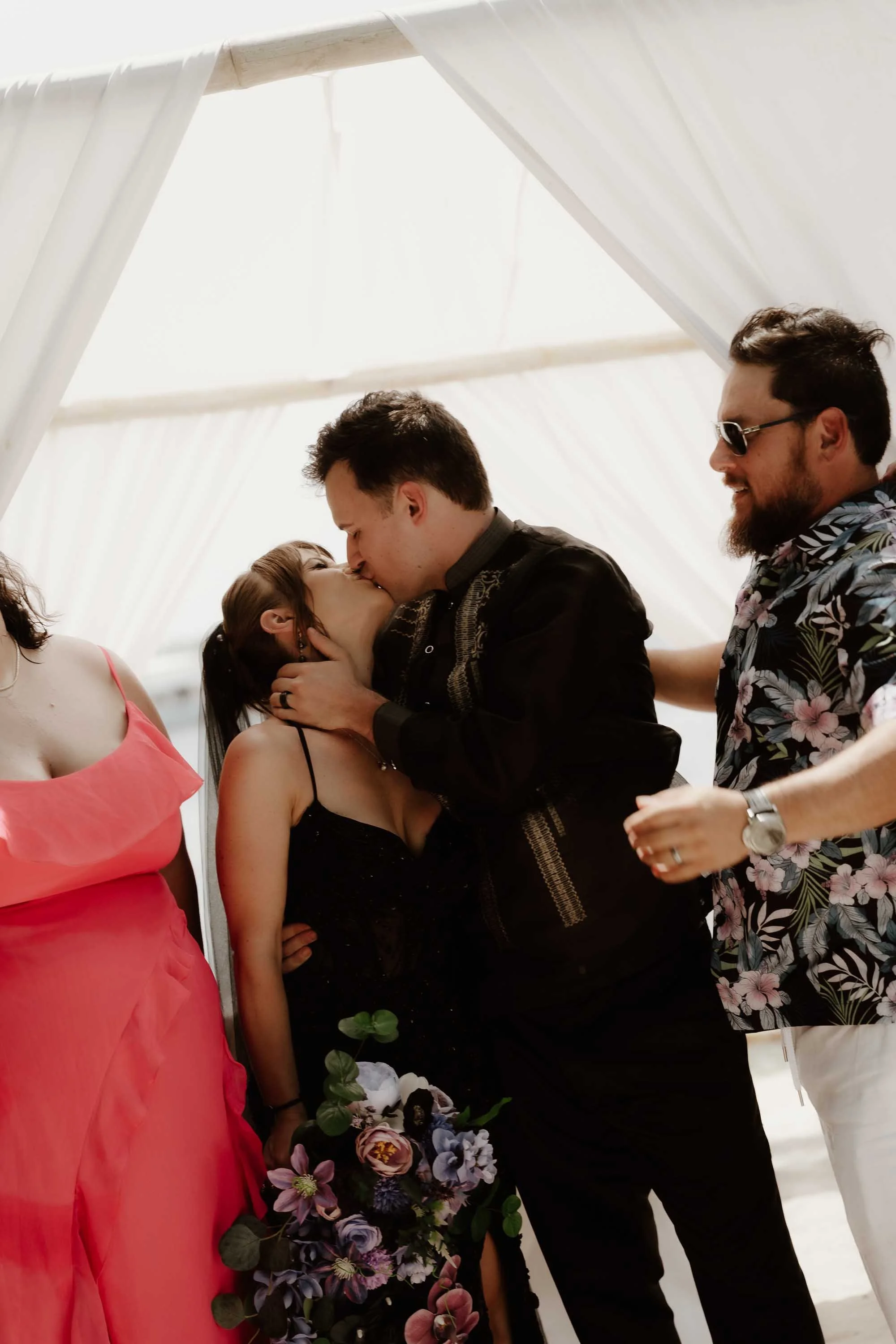 Group and family photo of Tori and Andrew while they kiss in the ceremony after the wedding