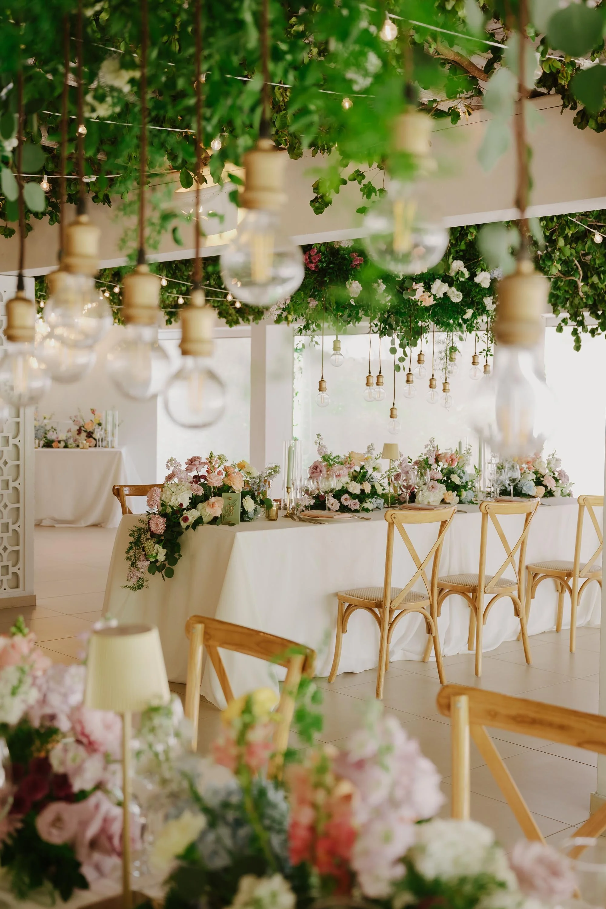 Elegant wedding reception table setup at Kukua Beach Club, featuring pastel floral centerpieces, wooden cross-back chairs, and hanging Edison bulbs intertwined with lush greenery.
