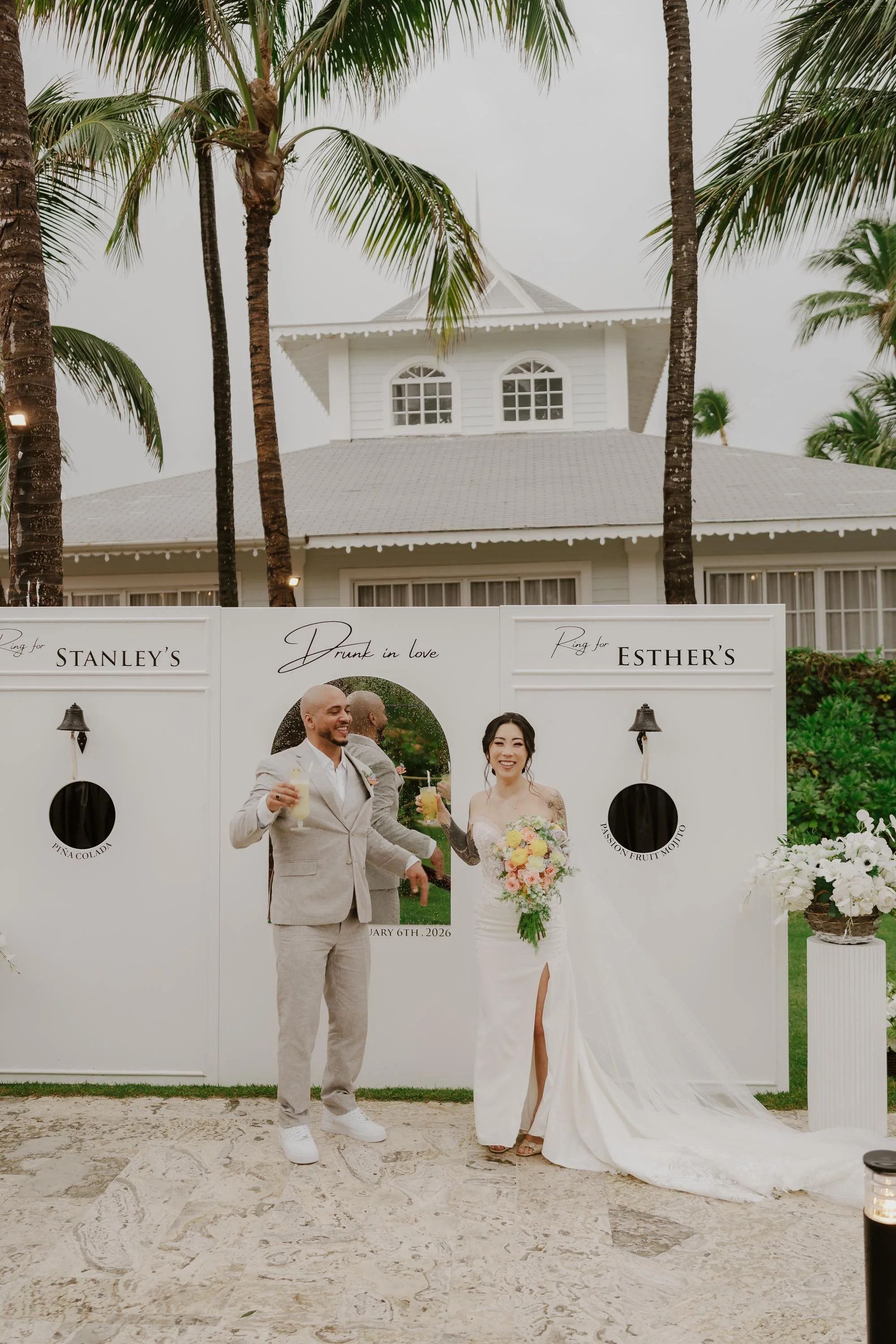 Bride and groom smiling and holding cocktails in front of a custom white "Ring Bar" setup at their wedding reception