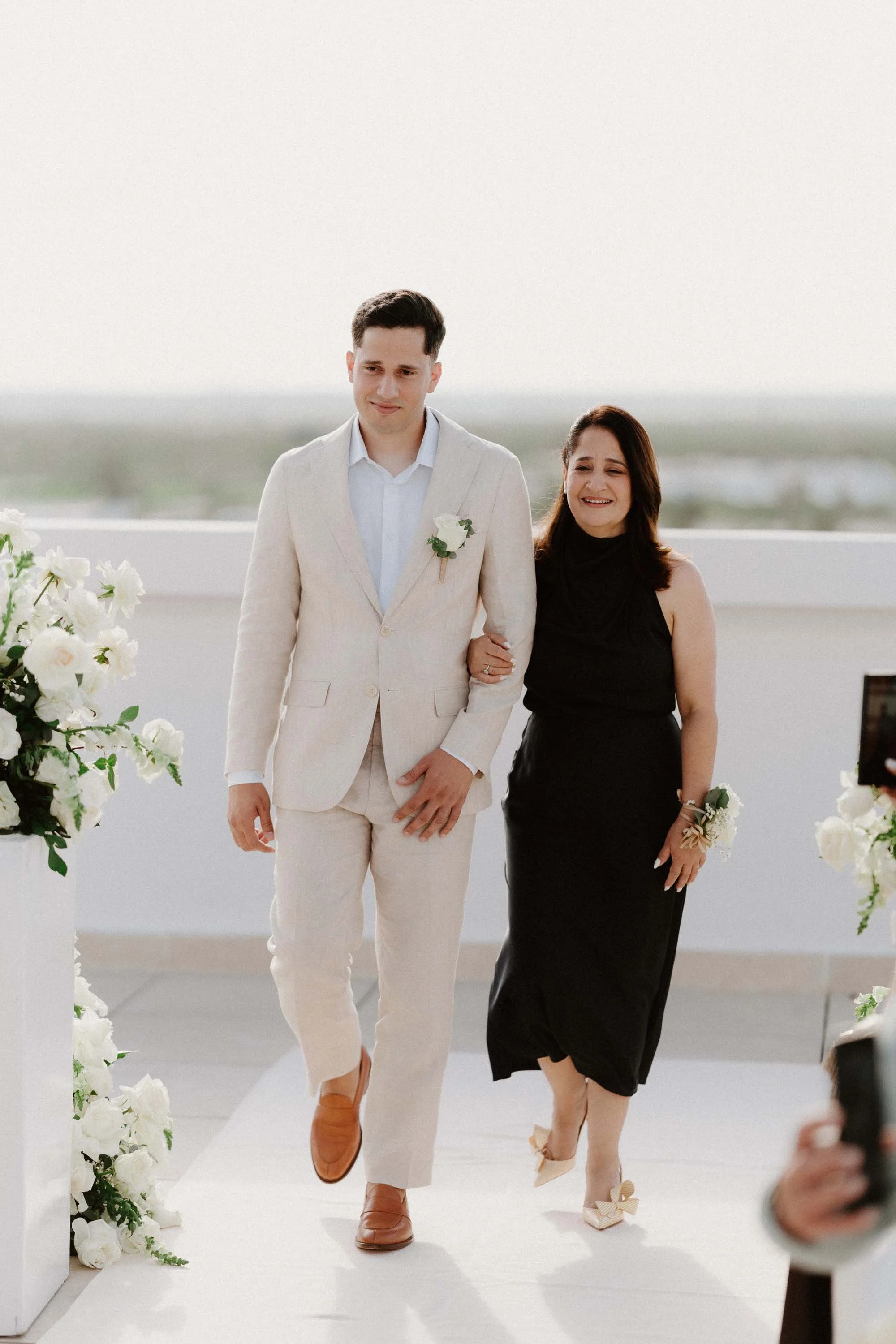 "A candid and emotional moment of the groom in a light linen suit walking down the white aisle arm-in-arm with his mother during a beautiful oceanfront wedding ceremony in Cap Cana Punta Cana."