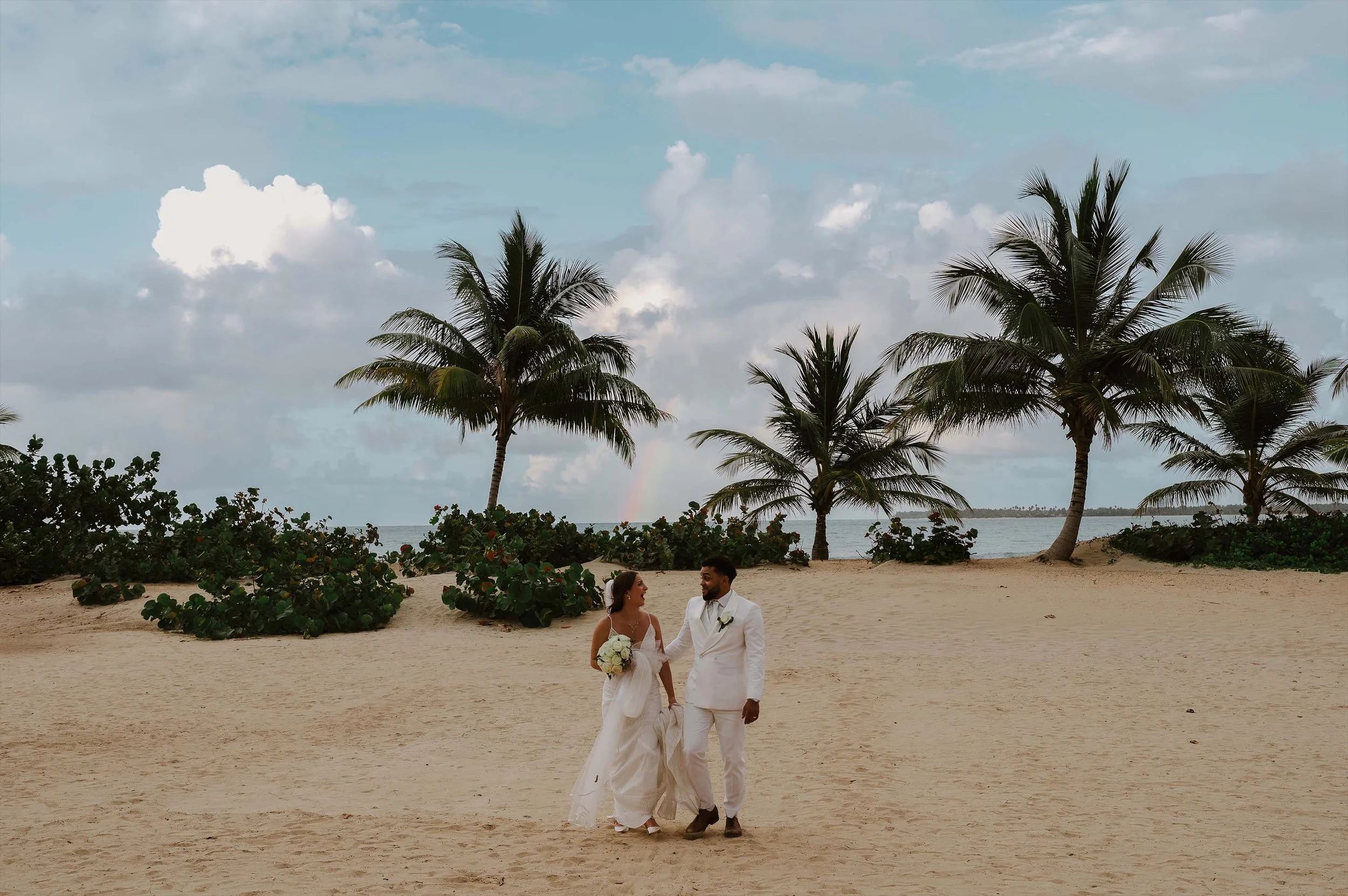 Couple walking towards the camera in the beach, at the background there is a rainbow and the beach