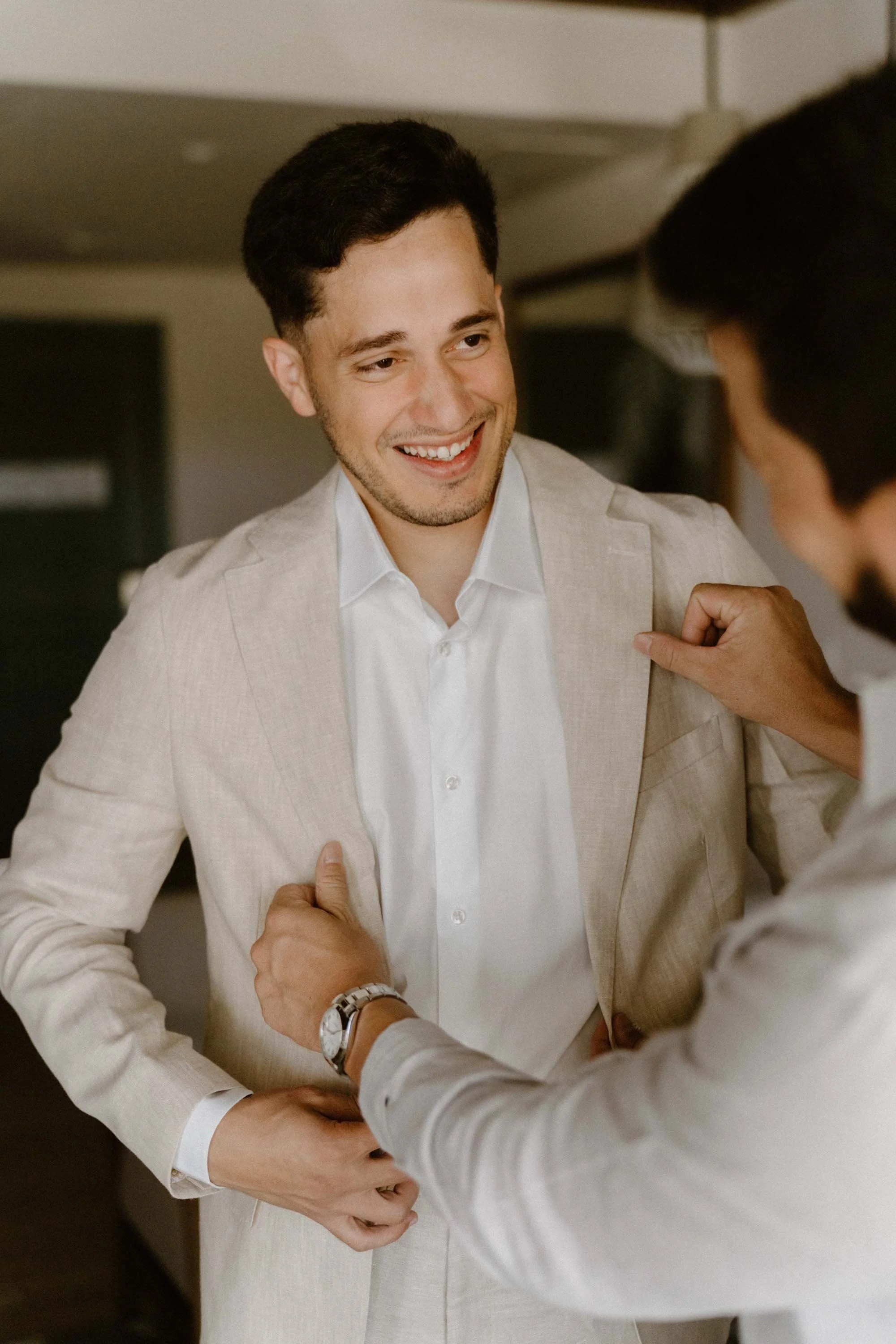 Candid close-up of a joyful groom smiling warmly as his best man helps adjust his light linen suit during the wedding morning prep in Cap Cana Punta Cana.